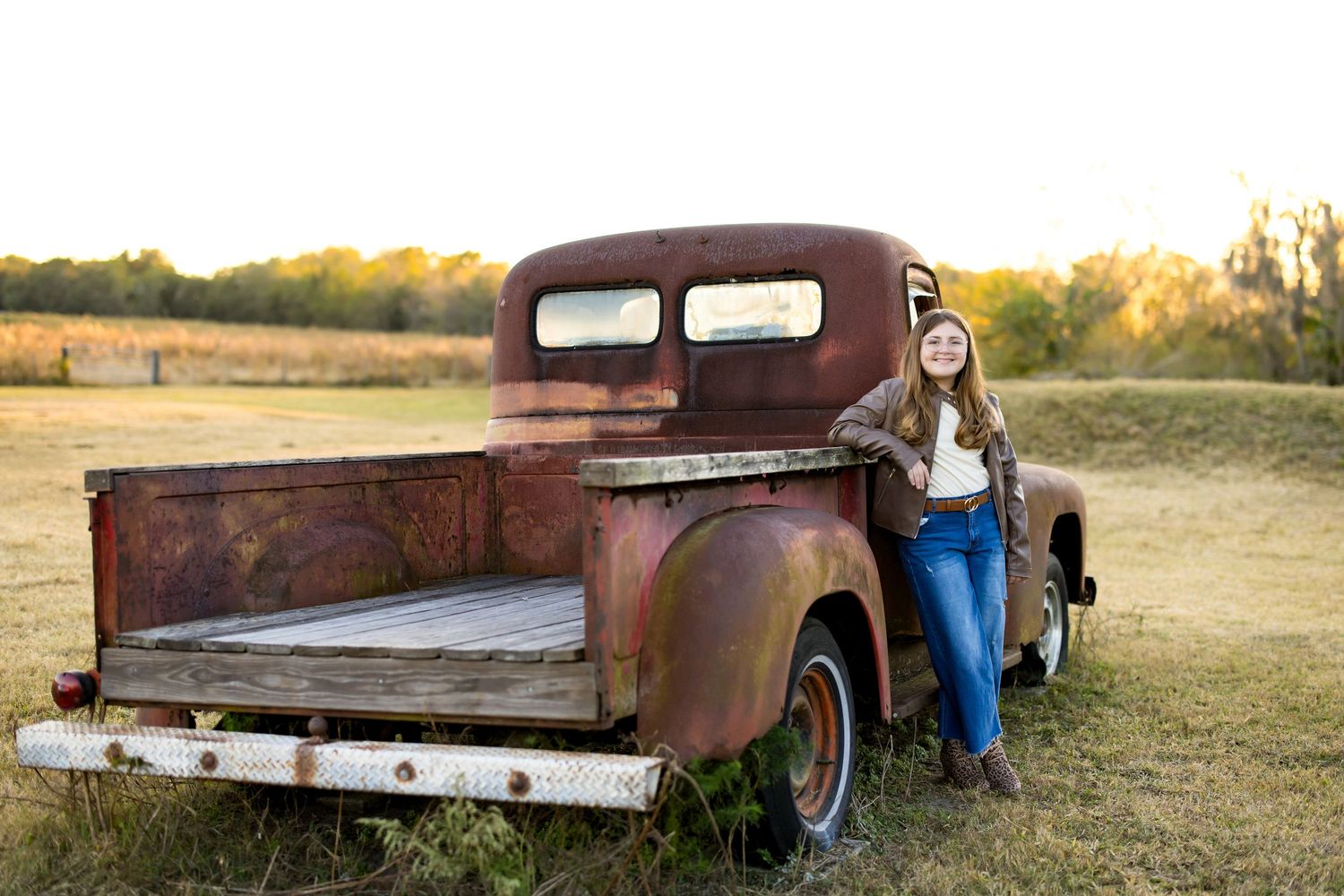 Young woman in jeans leaning against a rusty vintage pickup truck in a golden field at sunset.