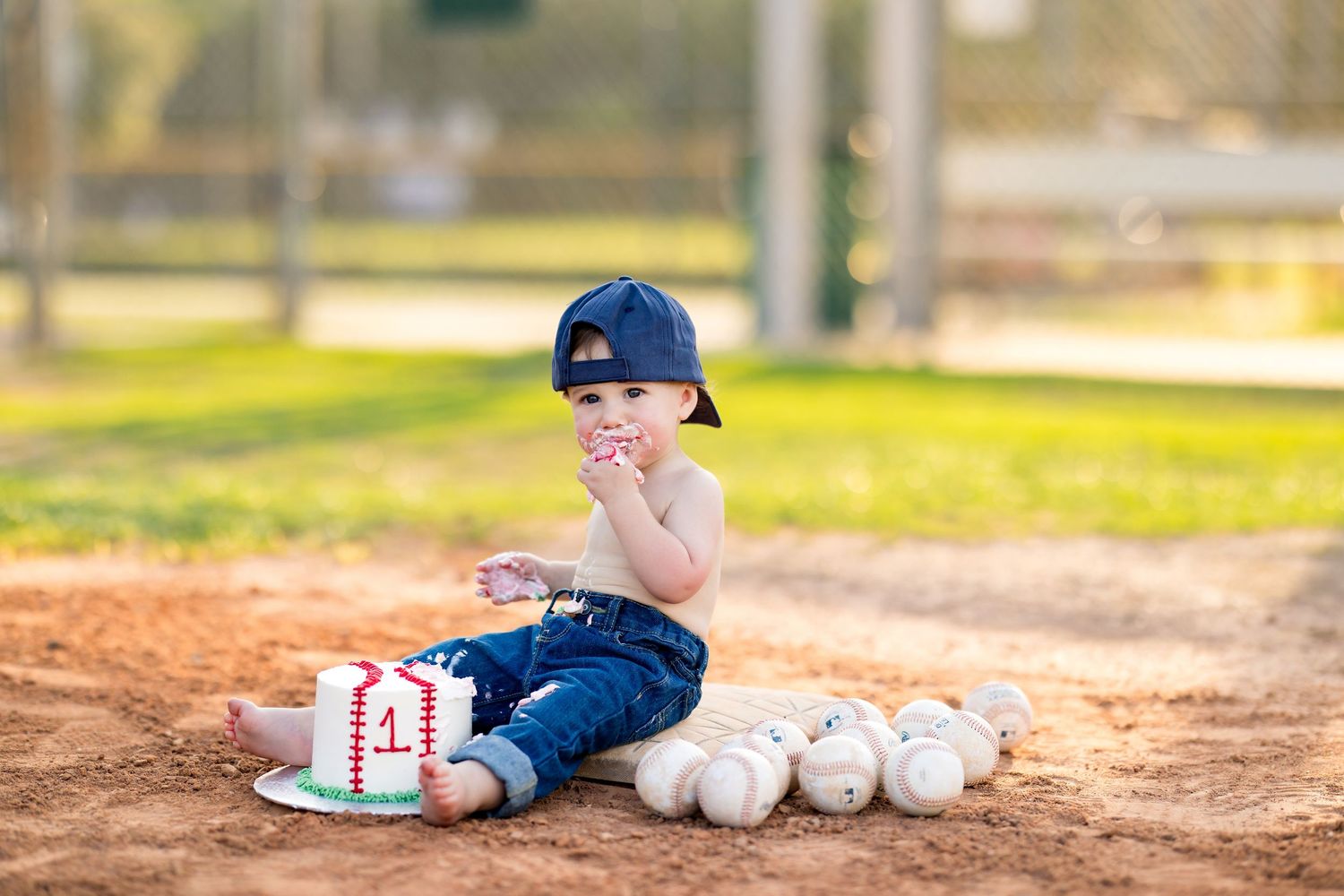 Baby boy in baseball cap sits on dirt field with cake and baseballs celebrating his 1st birthday.