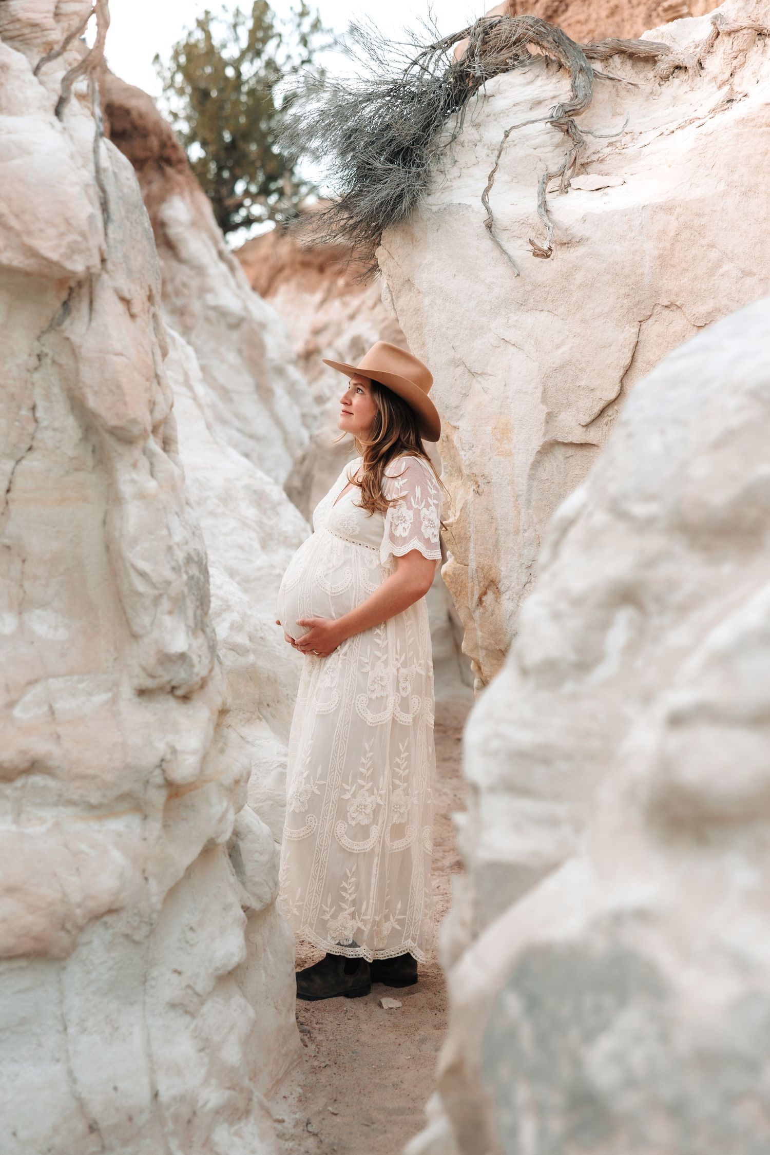 Pregnant woman in white lace dress and cowboy hat stands between white rock canyon walls.