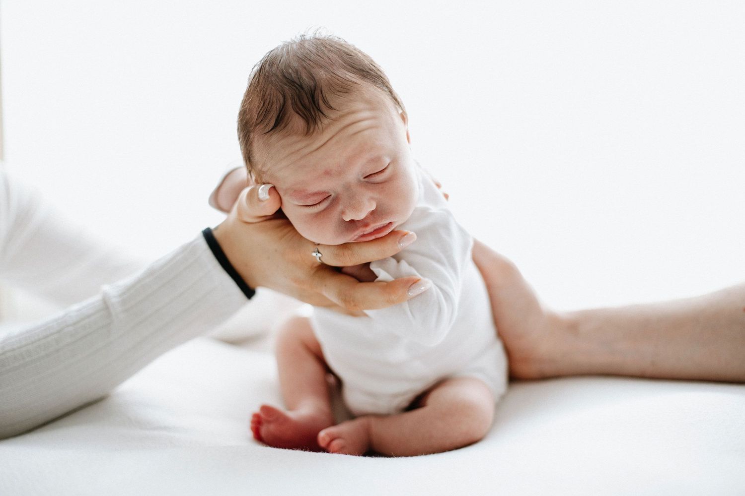 Newborn baby in white onesie being gently supported by both parent's hand on a soft white fabric covered beanbag.