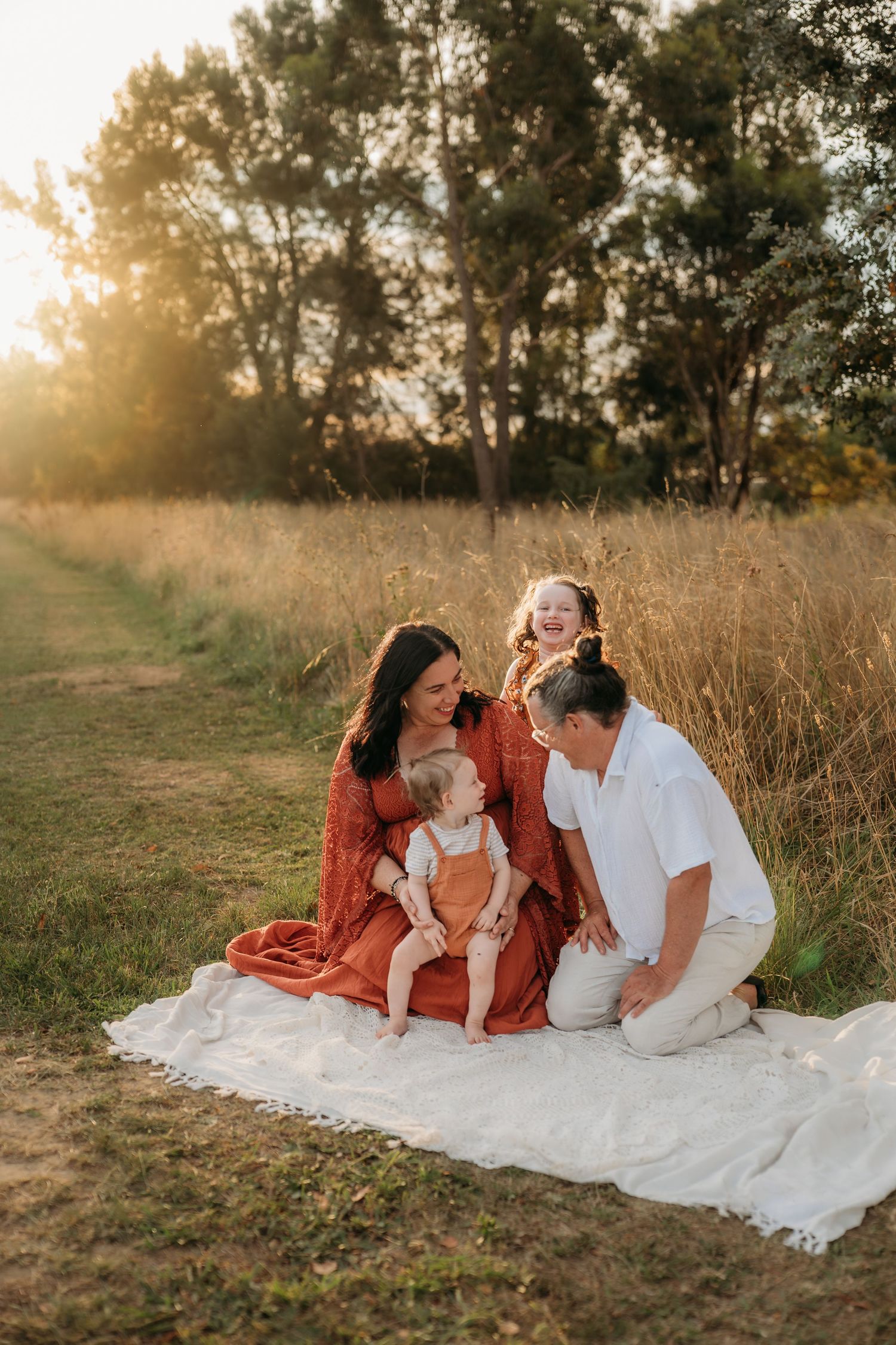 Family of four sitting on white blanket in sunny green field, mom laughing while dad cuddles two young boys.