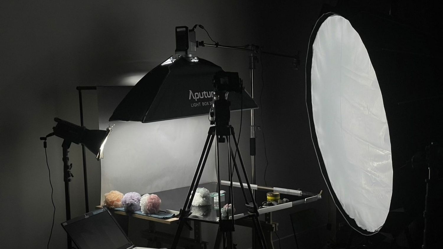 Professional product photography studio setup with softbox lights, tripod, and reflector on a dark background.