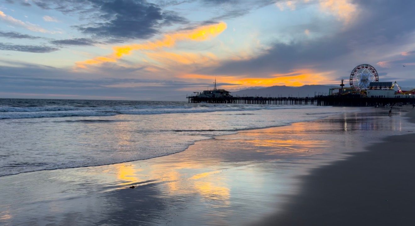 Stunning sunset at Santa Monica Pier with vibrant orange clouds reflecting on wet sand and Ferris wheel silhouette.