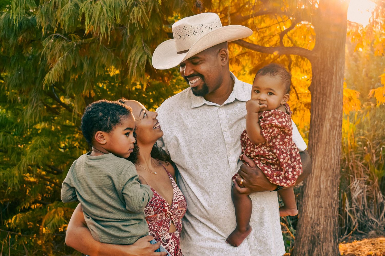 A joyful Black family with a father in a cowboy hat holding two young children outdoors in golden sunlight.