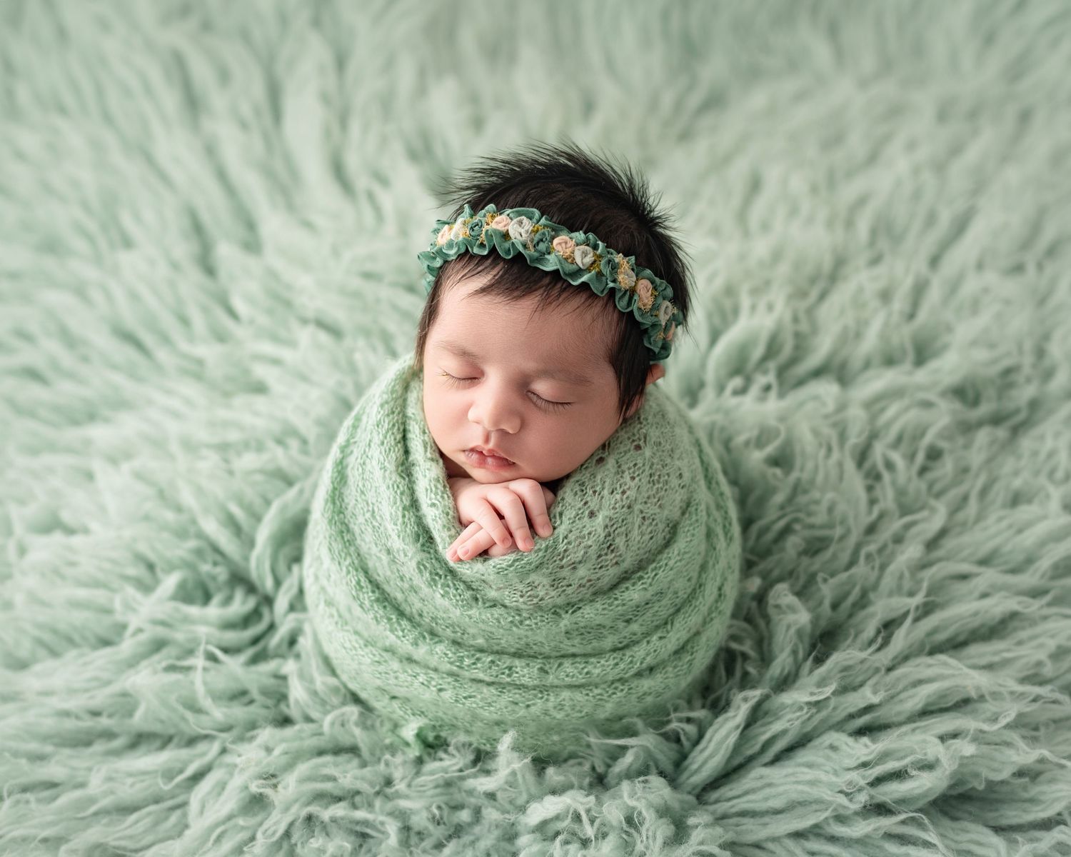 Sleeping newborn baby wrapped in green knit swaddle with floral headband on soft green fluffy rug.