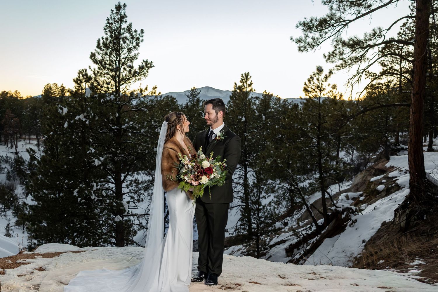 Bride and groom stand on snowy rocks surrounded by pine trees at sunset in a romantic winter wedding portrait.