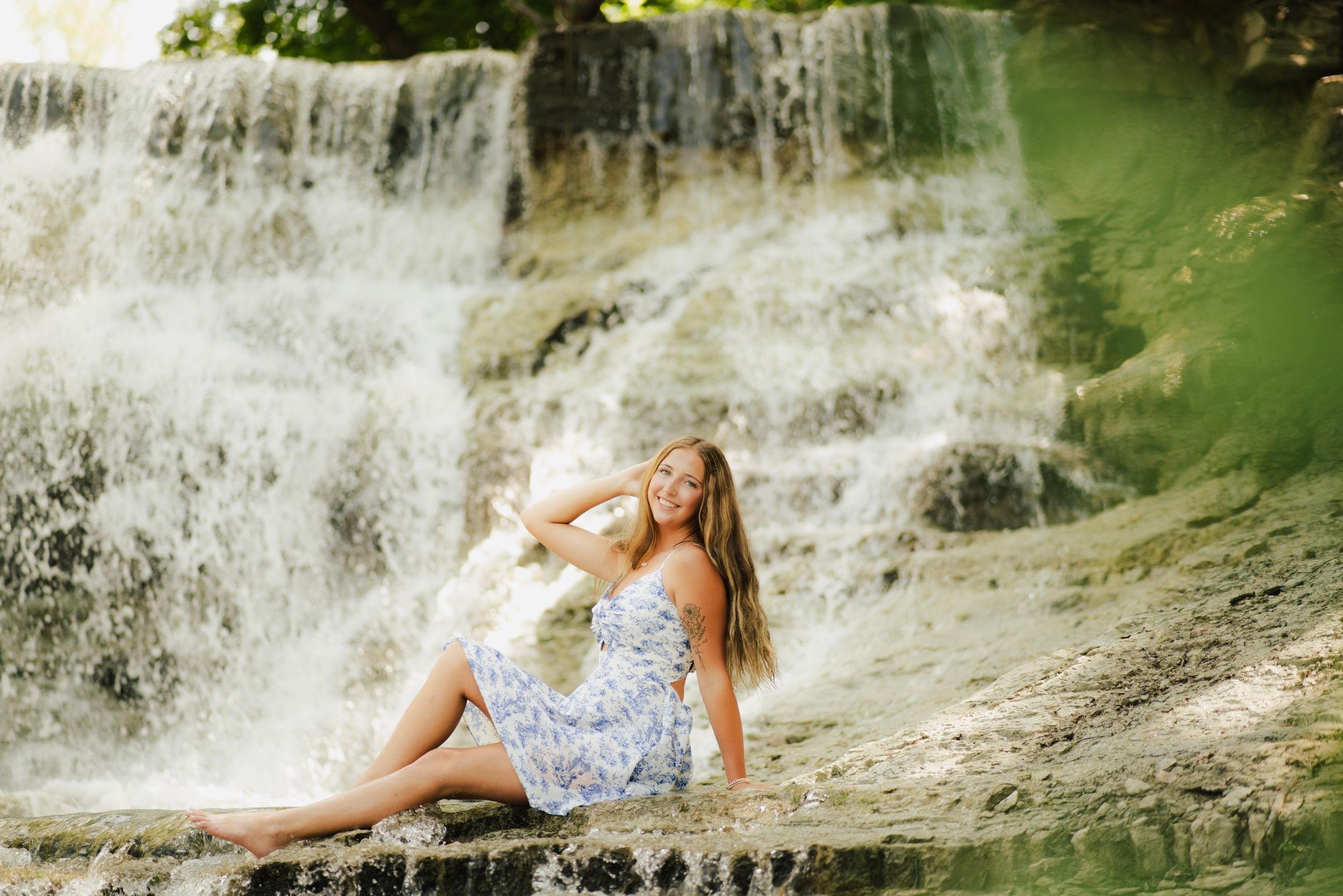 Young woman in floral dress sitting on rocks in front of a cascading waterfall outdoors.