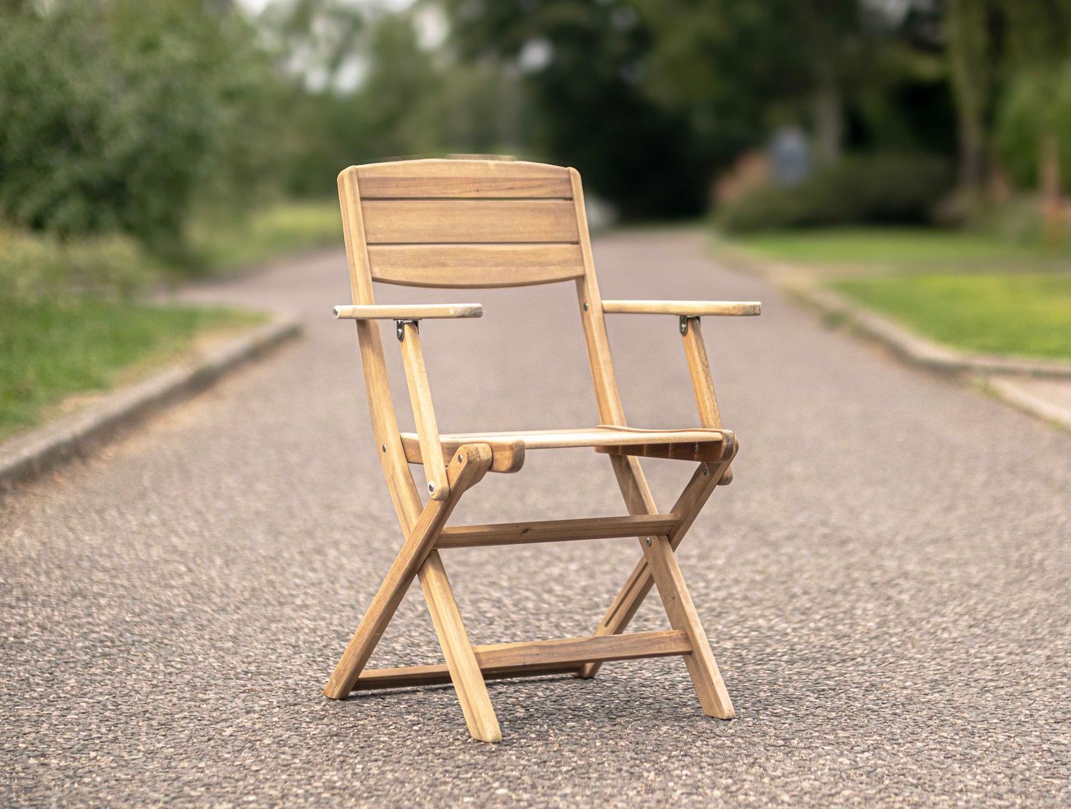 Folding teak wooden armchair placed on a paved path surrounded by lush green trees.