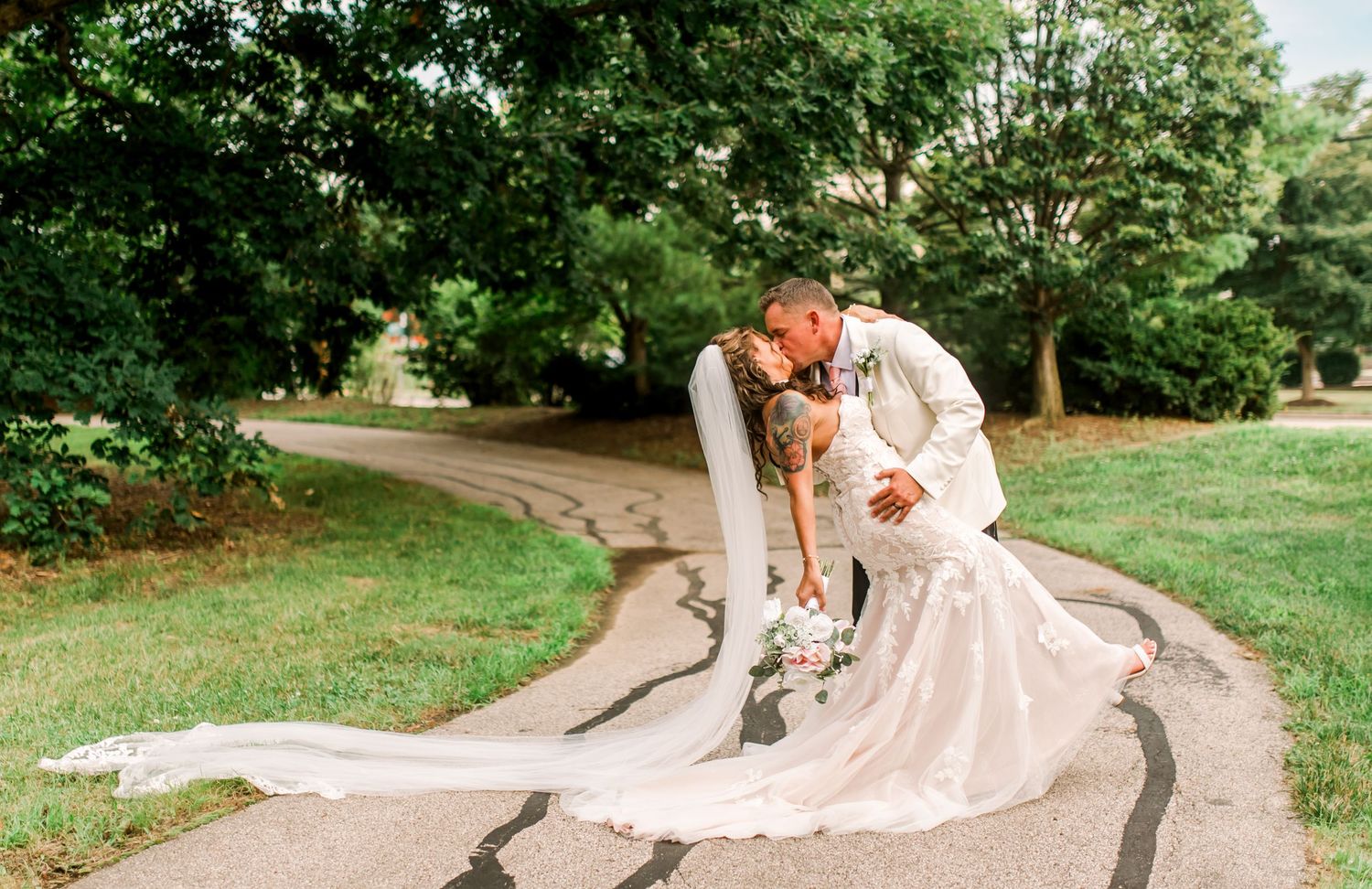 Bride and groom share a romantic dip kiss on a winding path surrounded by lush green trees on their wedding day.