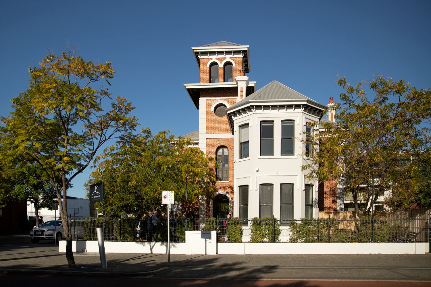 Historic Victorian brick mansion with tower, arched windows, and white fence surrounded by trees on a sunny day.