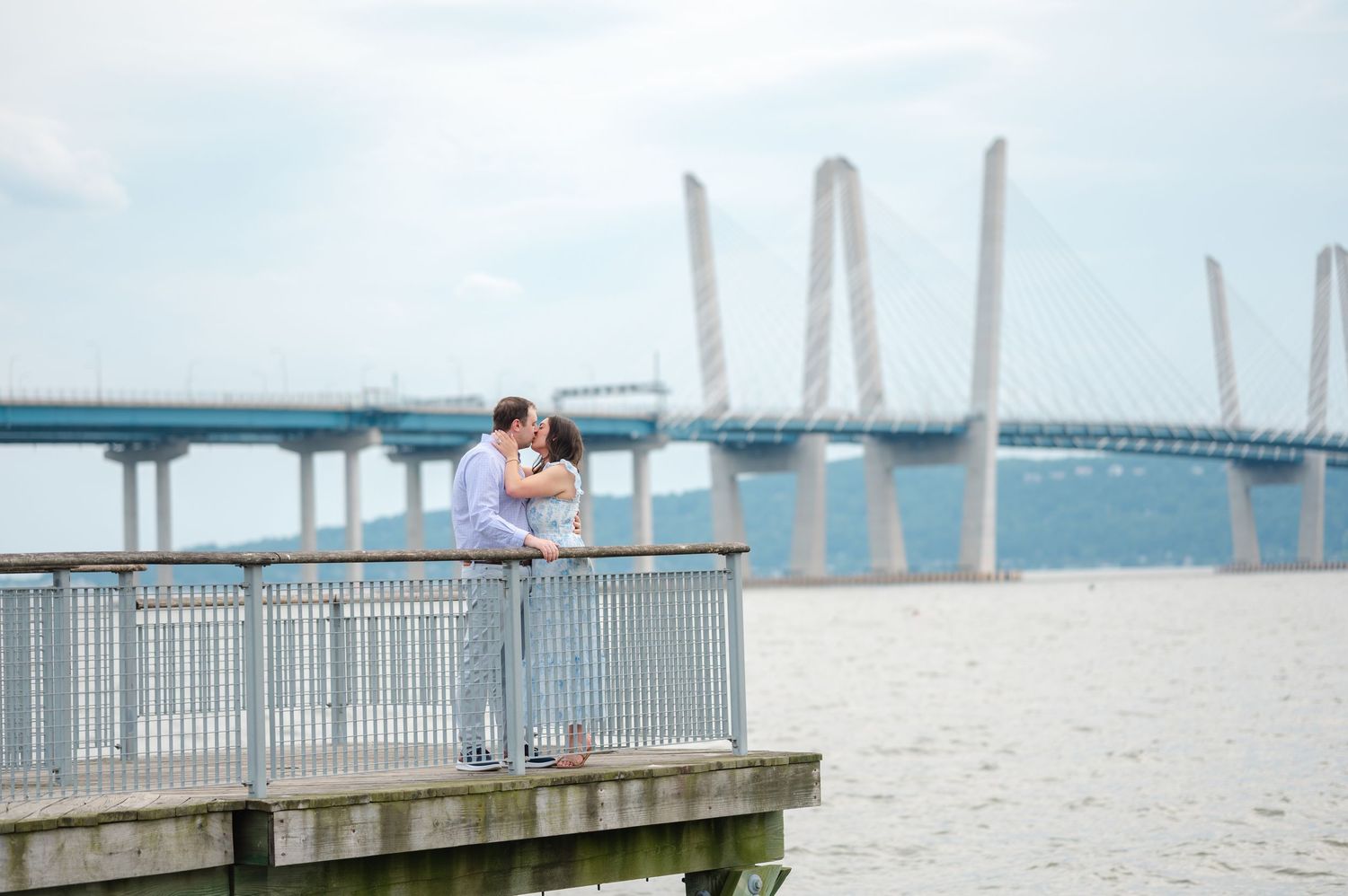 Couple kissing on a waterfront dock with a cable-stayed bridge in the background on a cloudy day.