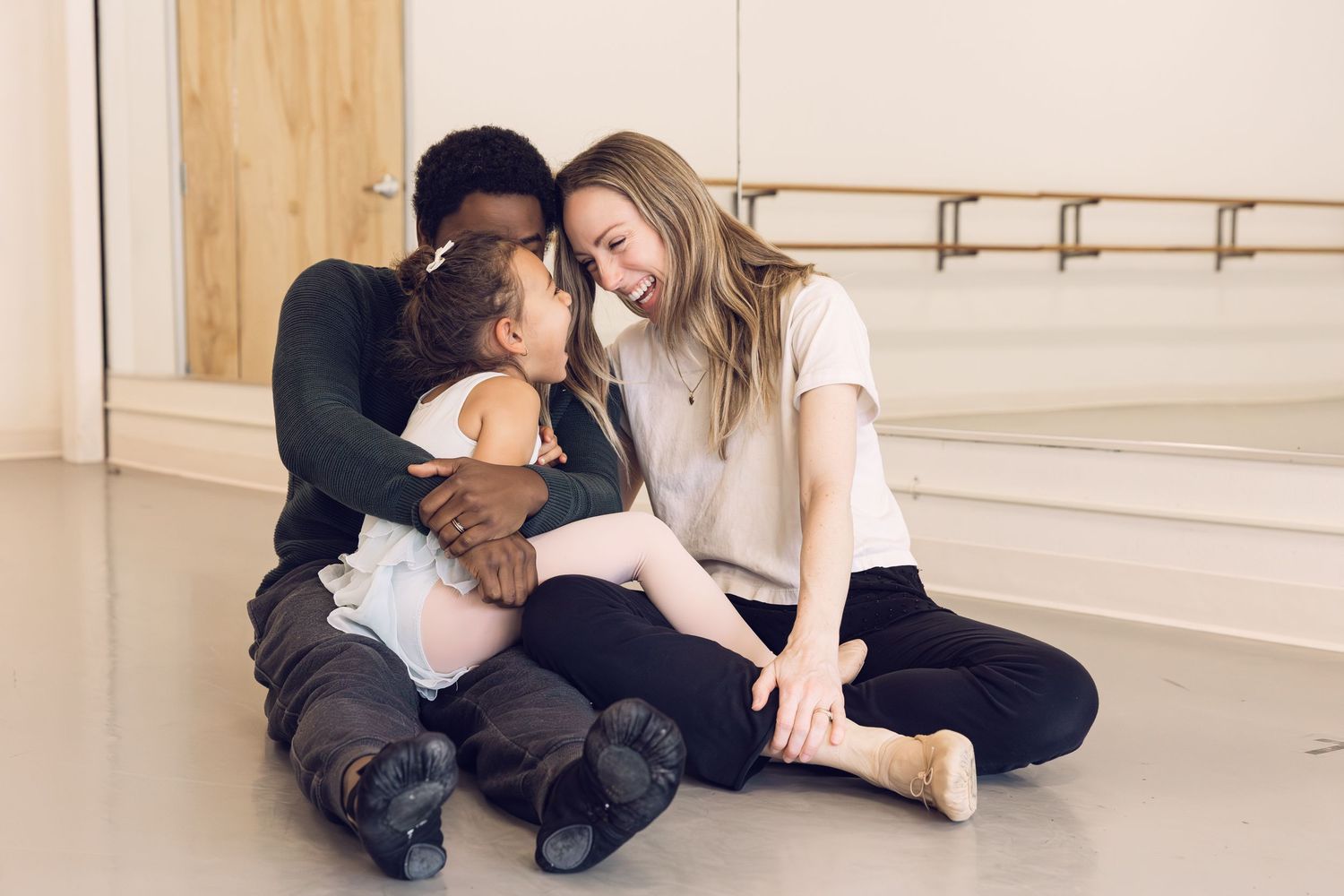 An interracial couple sharing a tender moment while embracing a young girl in a ballet studio.