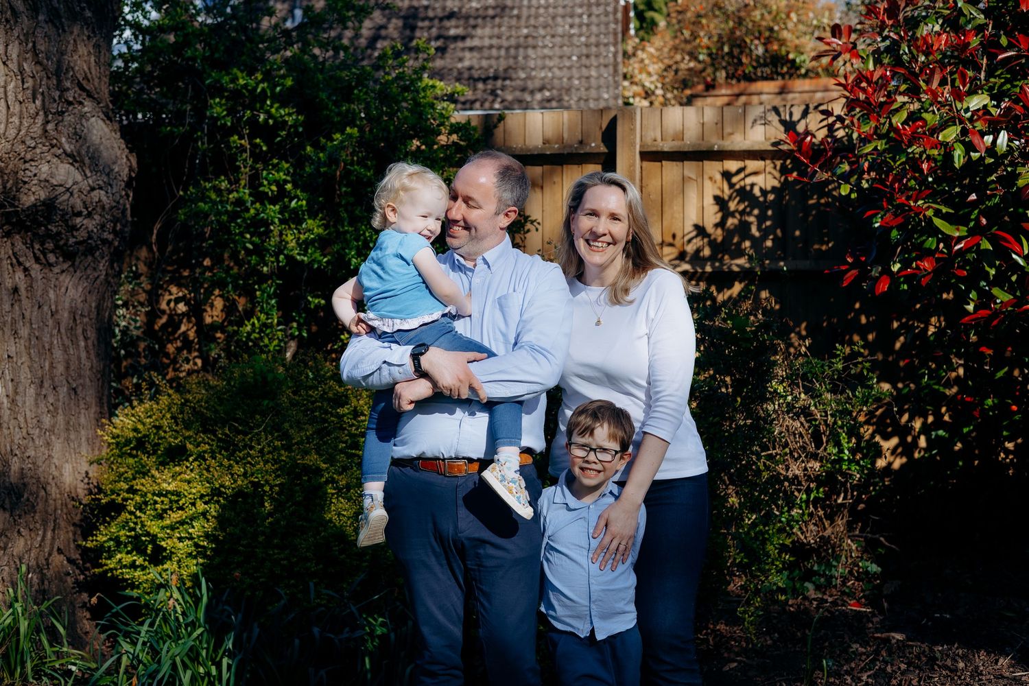 Happy family of four posing outdoors in a lush garden, parents holding young children while smiling.