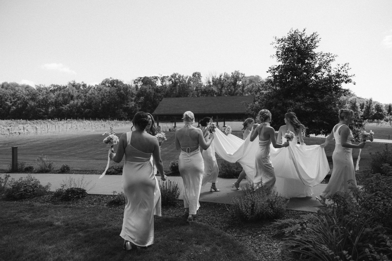 Black and white photo of bridesmaids in elegant gowns walking toward a Wisconsin rustic barn venue, carrying a wedding dress train in