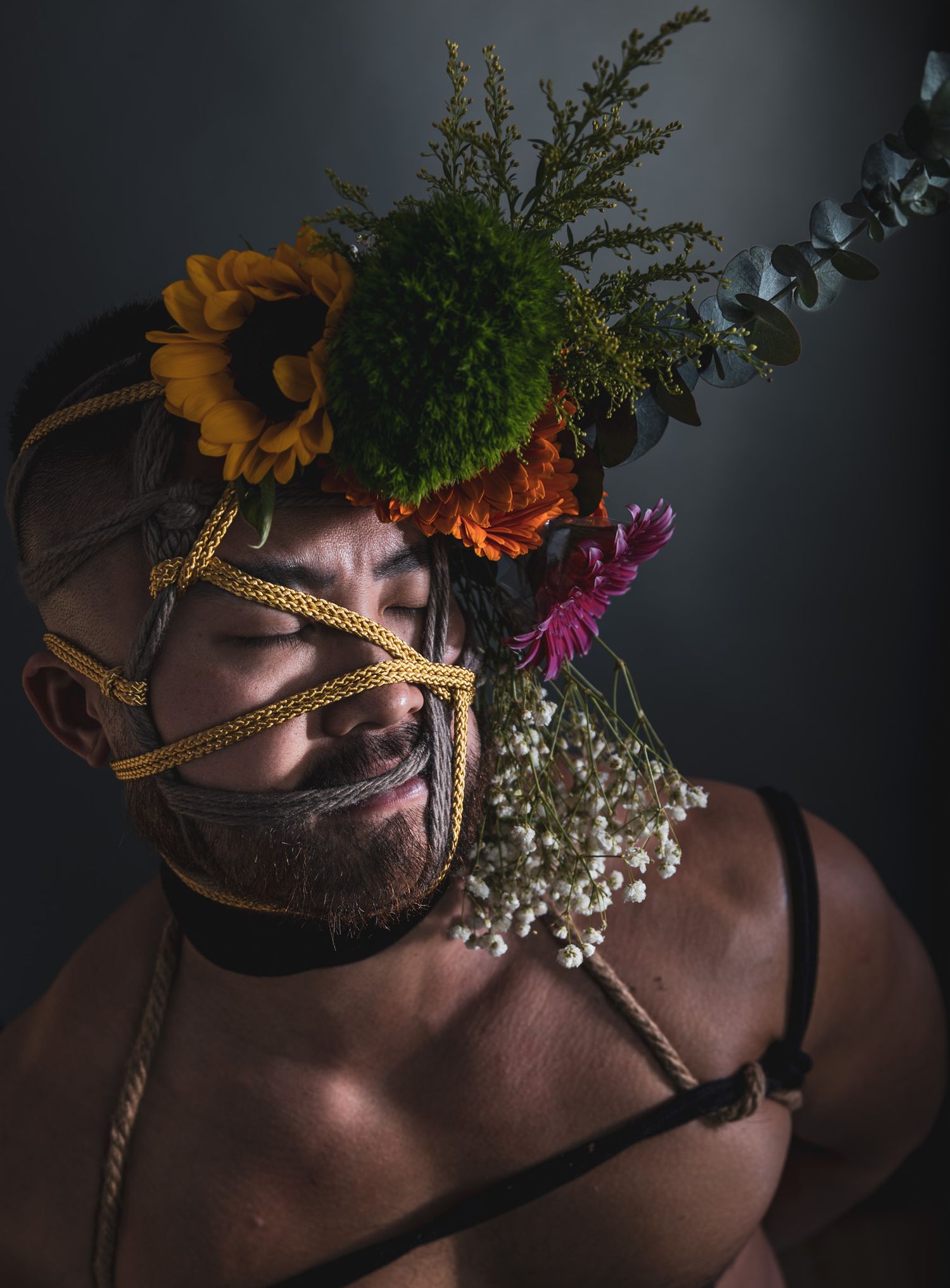 Bearded man with eyes closed wearing yellow rope harness and colorful floral crown with sunflowers and greenery.