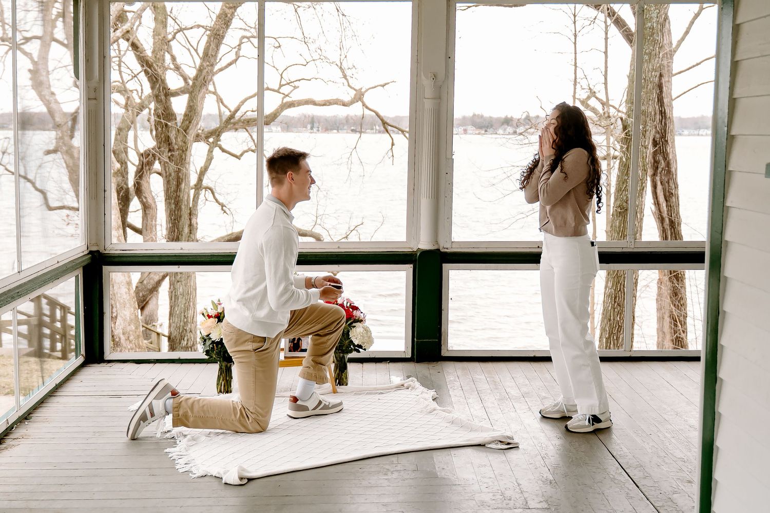 Man on one knee proposing to a woman on a sunlit porch with flowers and a white blanket nearby.