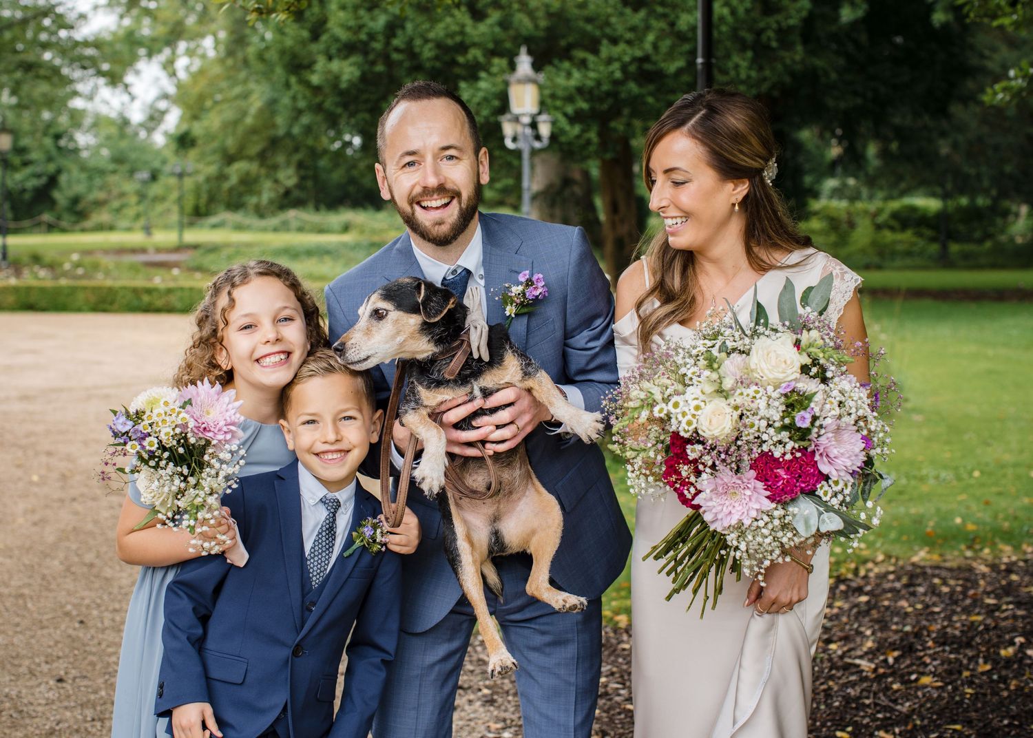 Wedding couple smiling with two children and a dog, bride holding floral bouquet, groom in blue suit, outdoors.