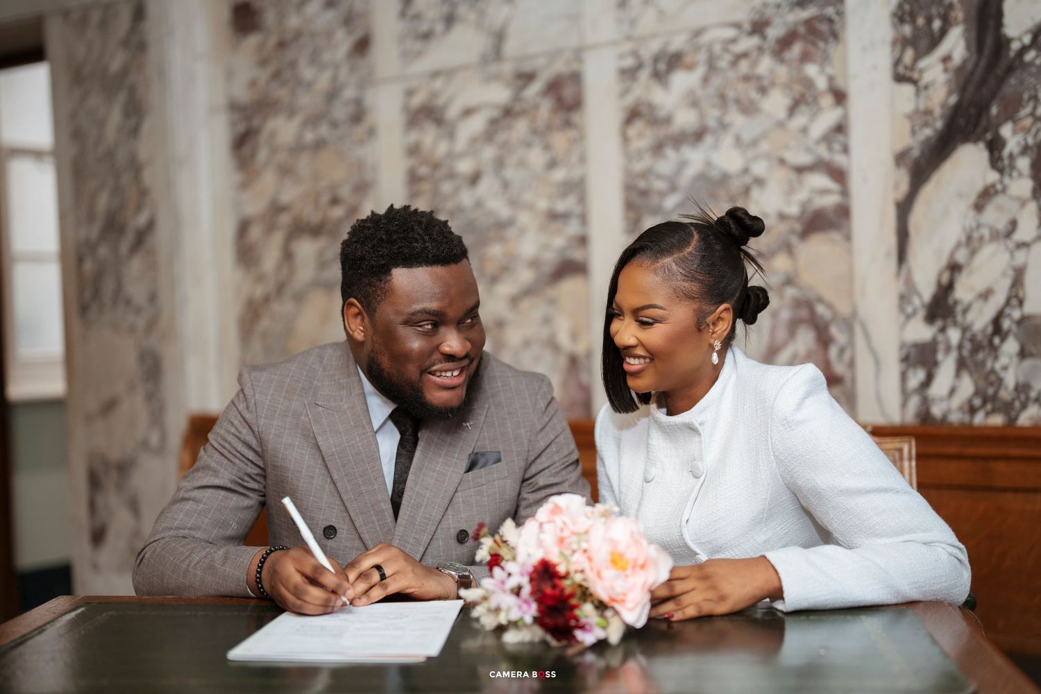 Happy couple signing wedding documents at a elegant venue, bride in white and groom in gray suit with floral bouquet.