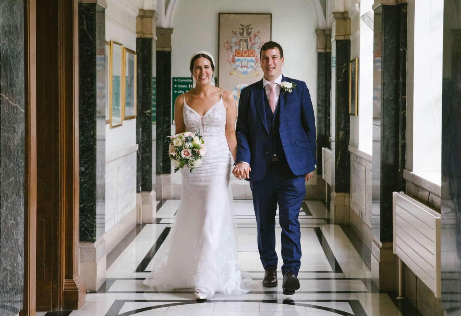 Bride and groom walking hand in hand through an elegant hallway with marble floors and natural light on their wedding day.