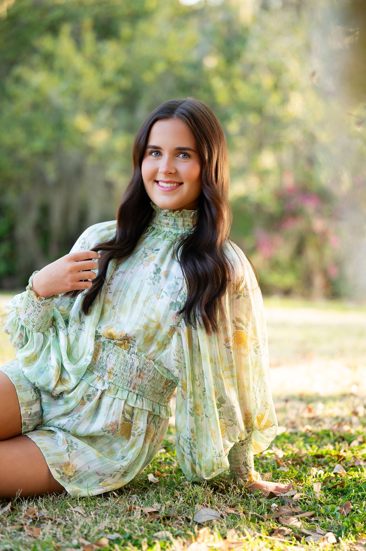 Young woman in green floral dress sitting on grass outdoors, smiling, with lush green trees in background.