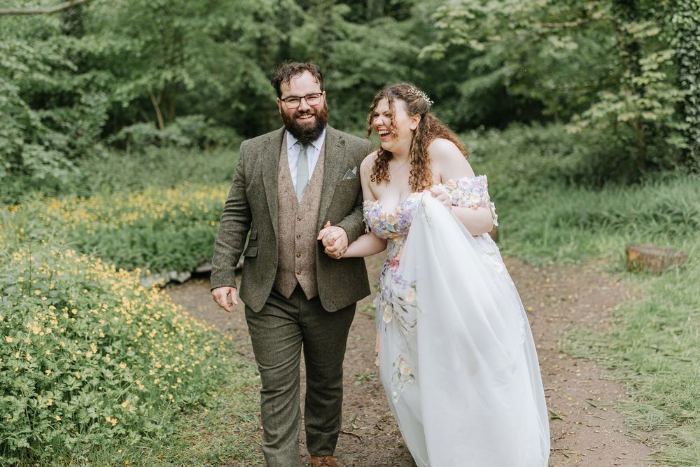 Bride in floral gown and groom in brown suit walk down aisle at outdoor woodland wedding ceremony.