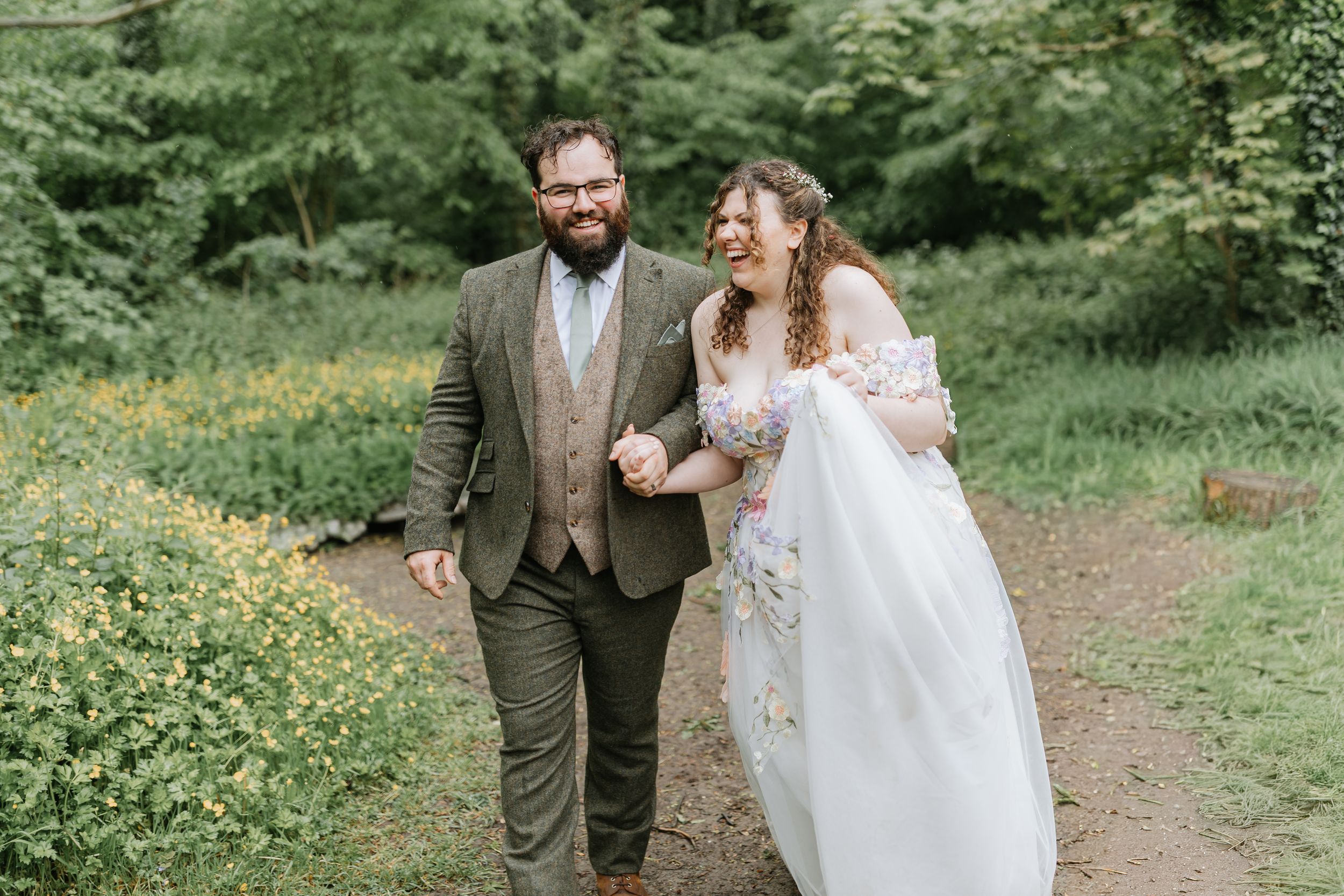 Bride in floral gown and groom in brown suit walk down aisle at outdoor woodland wedding ceremony.