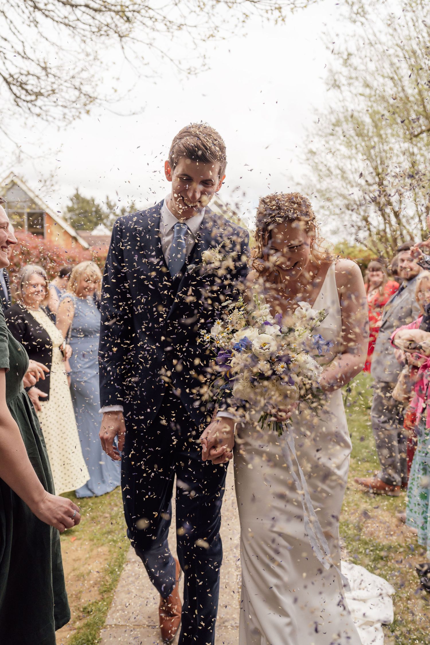 Bride and groom walk through confetti shower surrounded by wedding guests outdoors on their special day.