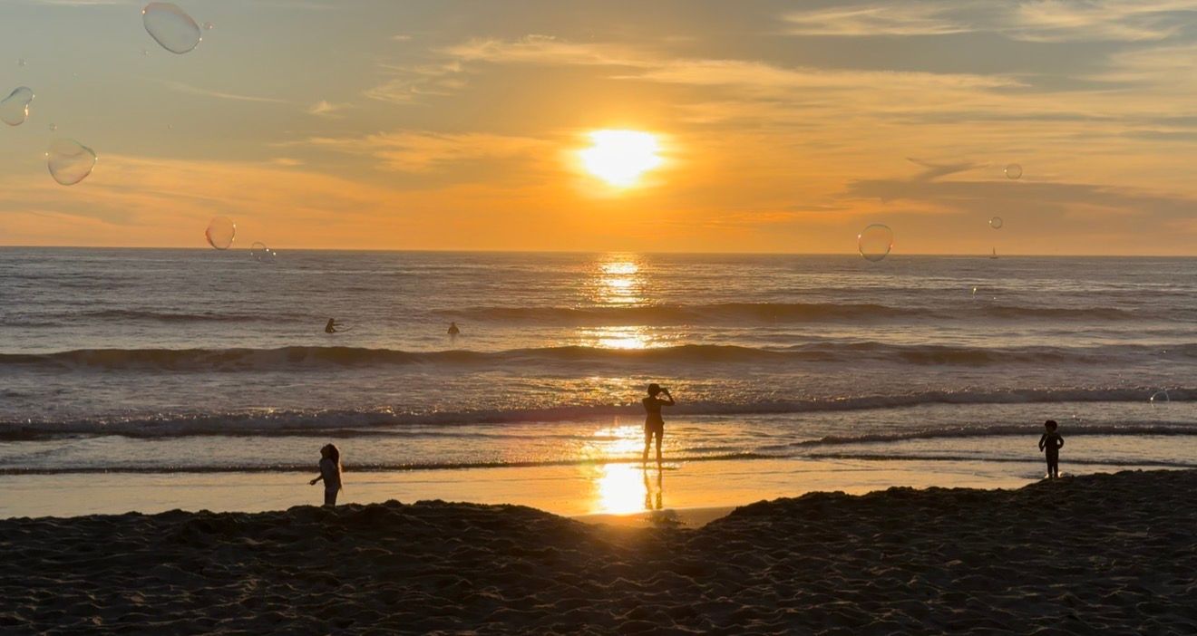 Silhouetted figures stand on a beach at sunset with golden light reflecting off ocean waves and dramatic clouds.