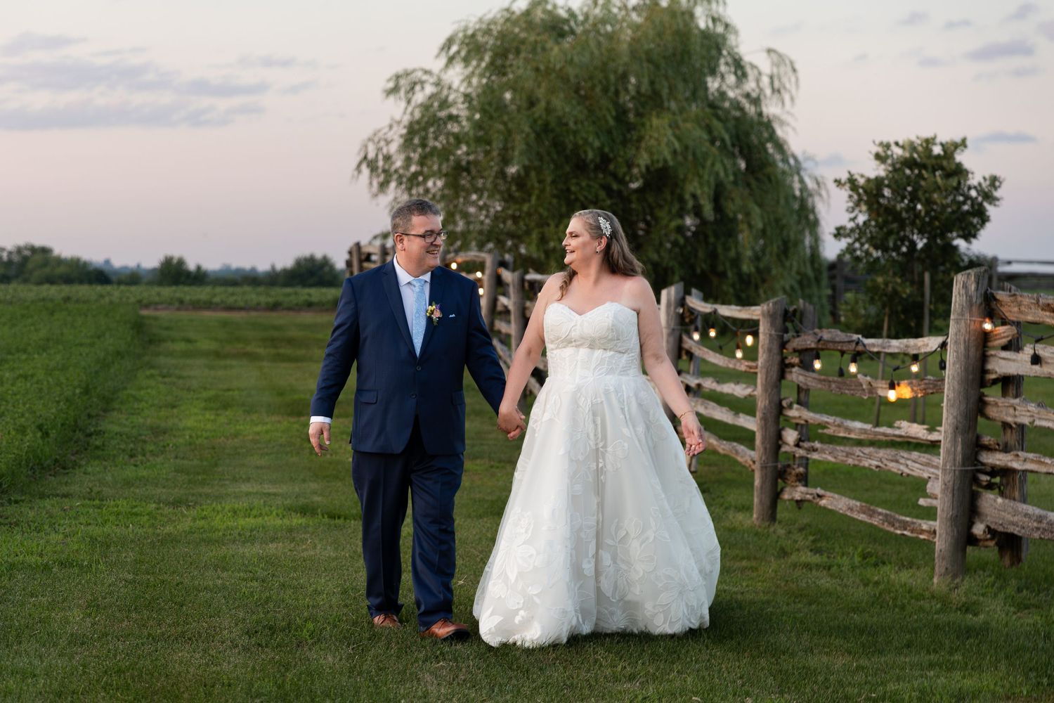 Bride & groom walking on a green field at dusk, with string lights and a wooden fence at barn swallow fields in Guelph, Ontario.
