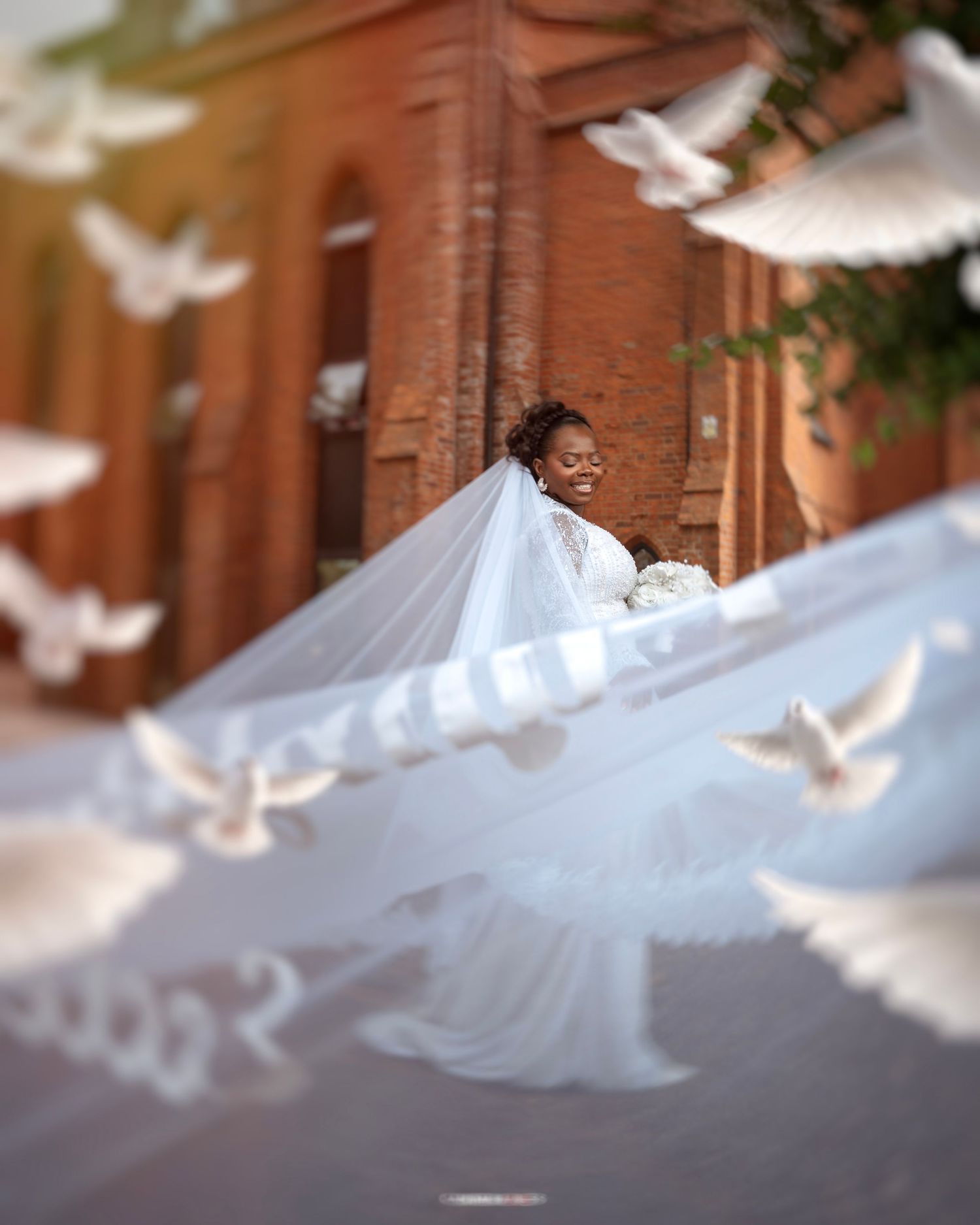 Bride in white gown and veil surrounded by flying white doves near a red brick building on her wedding day.