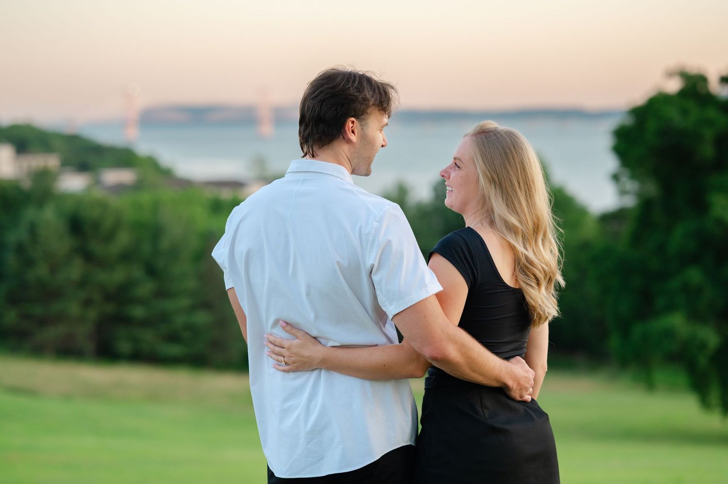 Man kneeling on one knee proposing to woman in black dress on grassy hill at sunset near waterfront.