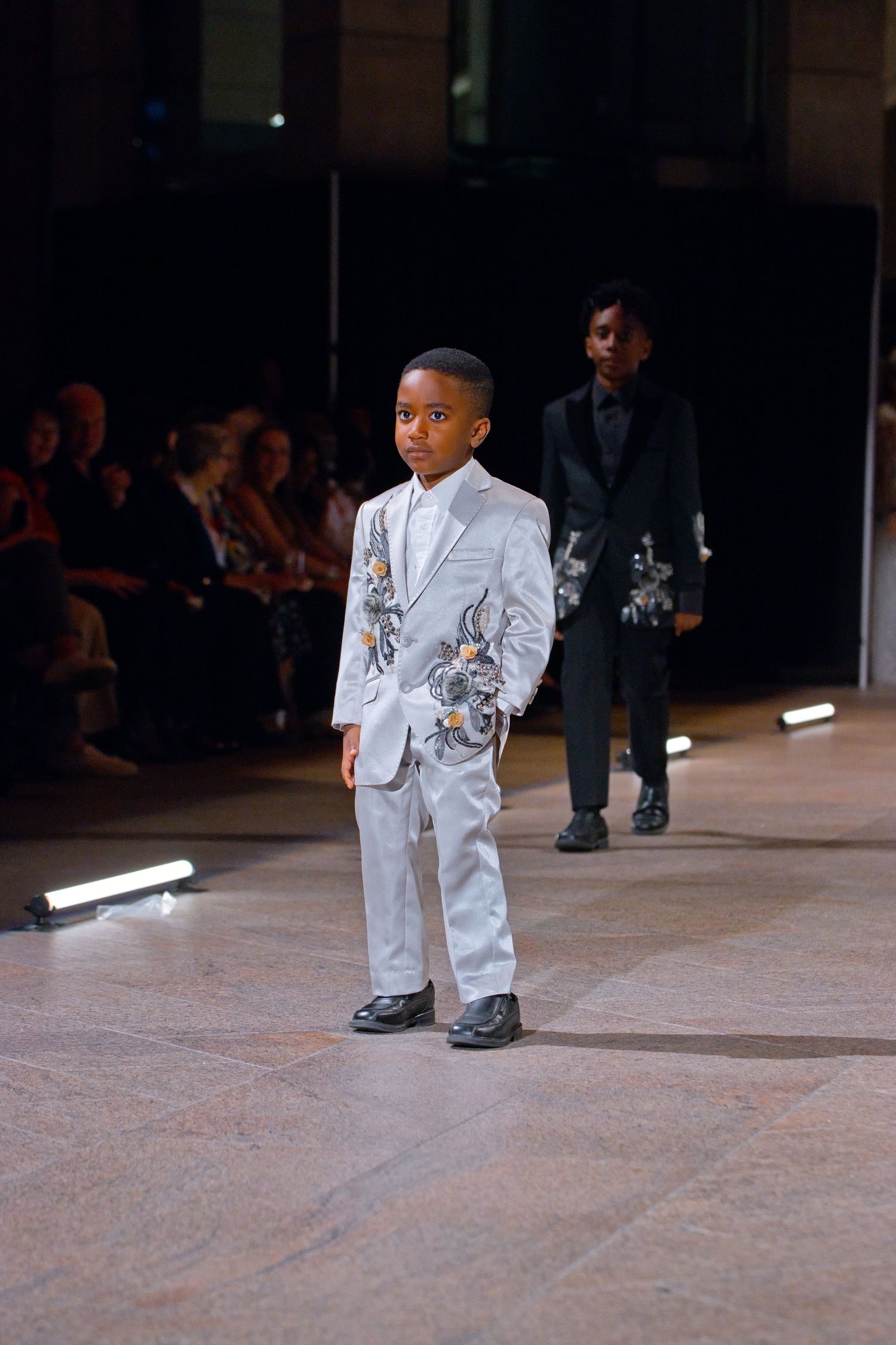 Young boy model walks runway in silver embroidered suit at fashion show, with another child model in black behind him.