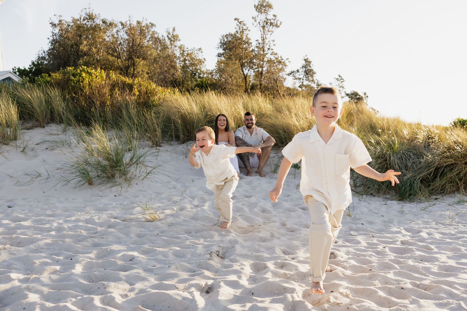 Two happy children run on sandy beach dunes while parents relax in background on sunny day.