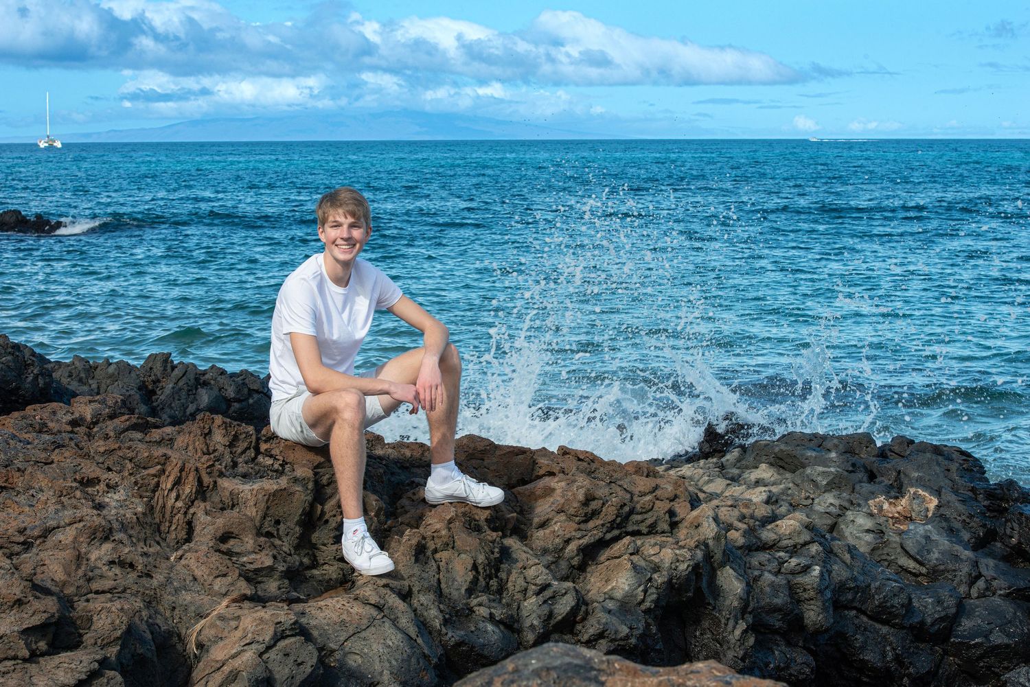 Young man in white shirt and shorts sitting on volcanic rocks by the ocean with waves splashing behind him.