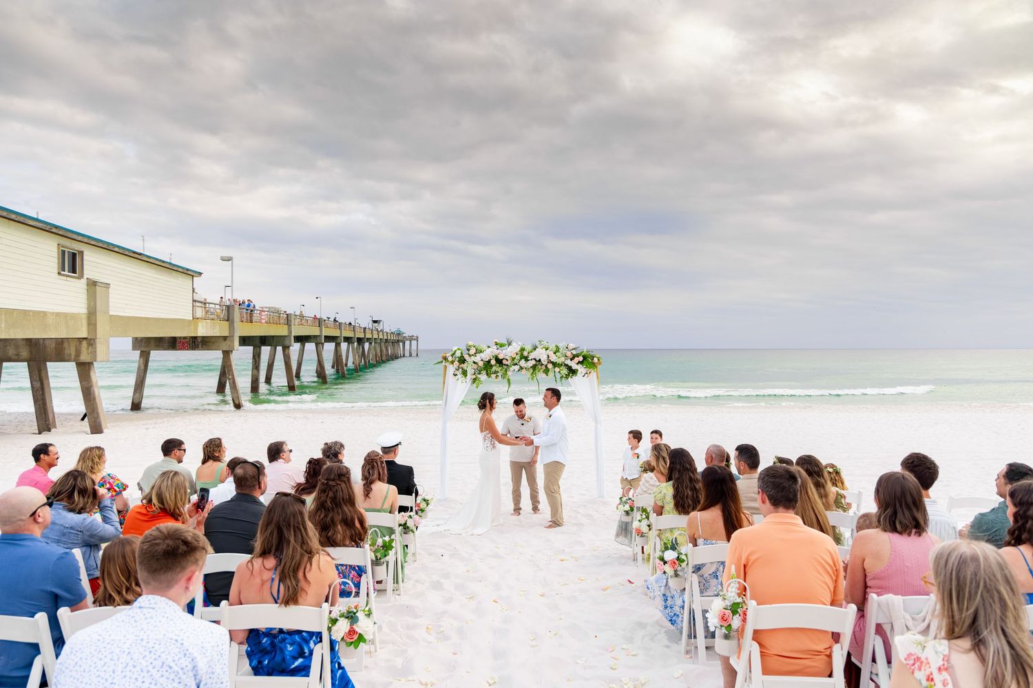 Bride and groom exchange rings on a beach wedding ceremony officiated by a smiling man in a white shirt.