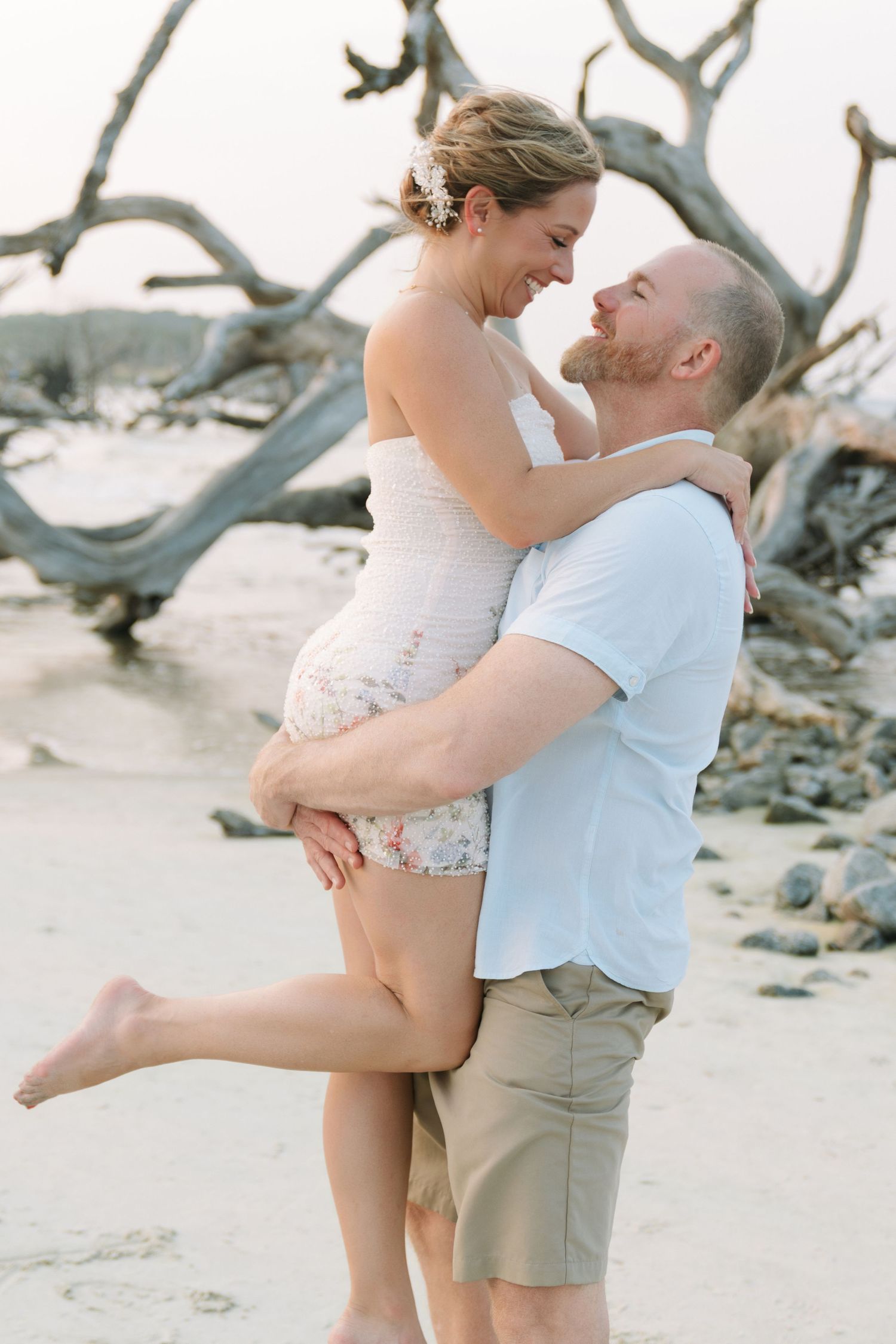 Bo lifts Rachel on the beach during their playful engagement session at Driftwood Beach