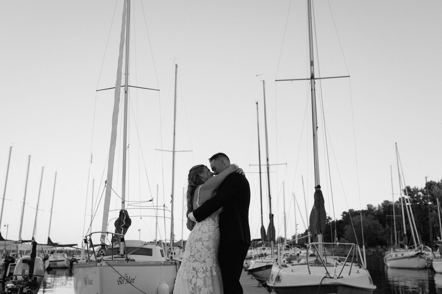 Black and white photo of a couple embracing at a marina with sailboat masts rising behind them.