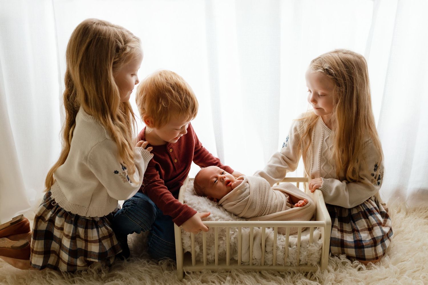 Three young siblings gather around a newborn baby in a small crib on a soft white rug near a bright window.