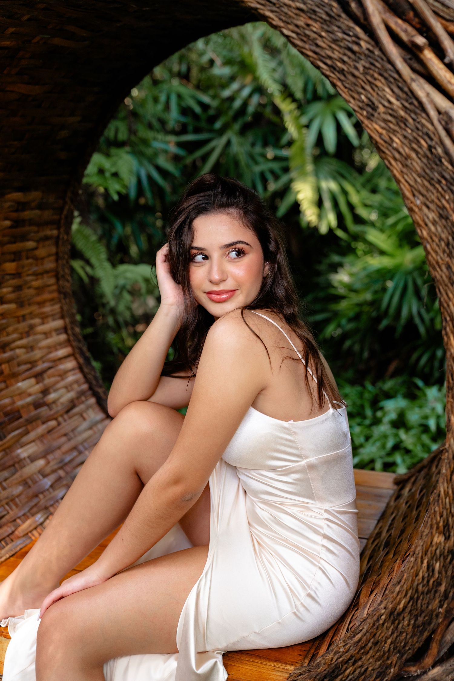 Young woman in white satin slip dress sitting in wicker arch surrounded by lush tropical greenery.