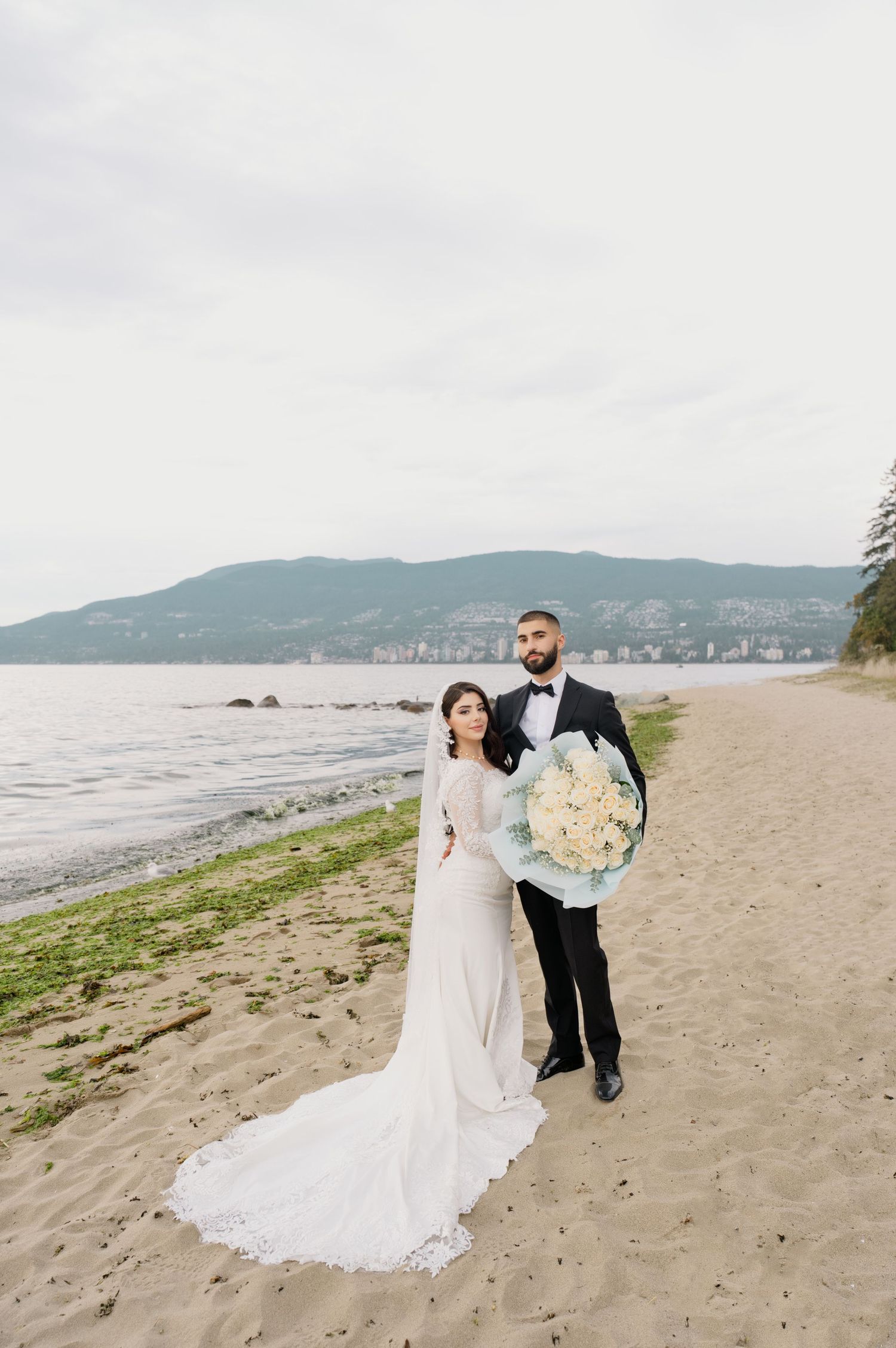 Bride in a lace wedding gown and groom in a black tuxedo holding a white bouquet pose on a sandy beach with mountains in the distance.