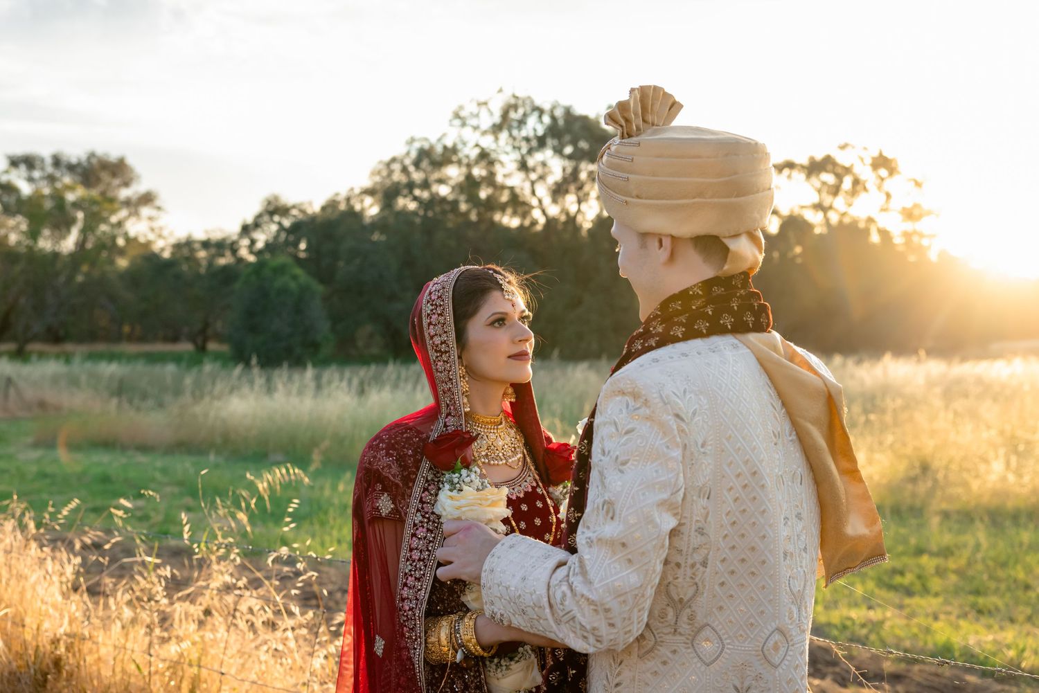 Bride in red lehenga and groom in ivory sherwani share a golden sunset moment at an outdoor Indian wedding.