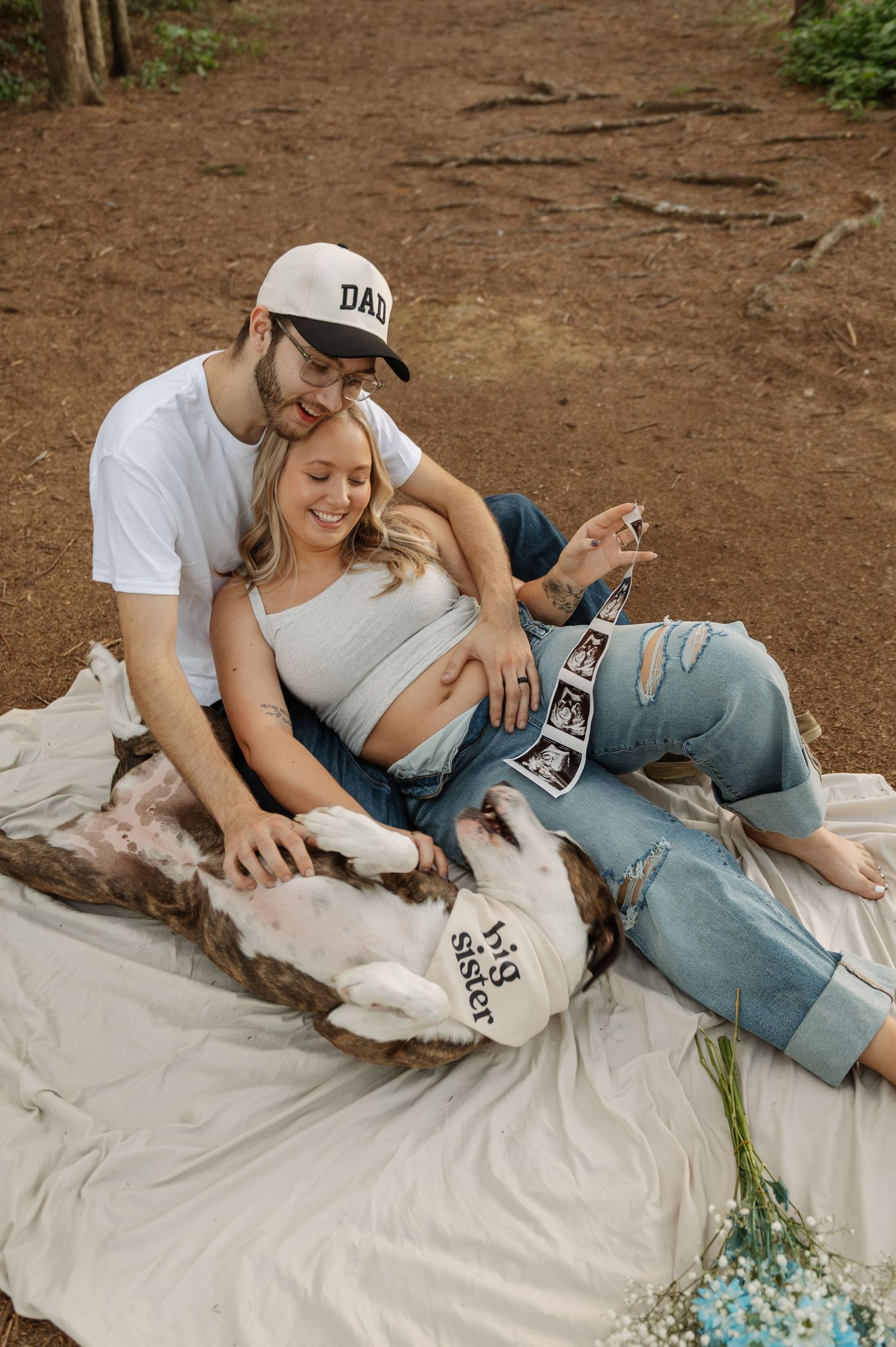 Pregnant couple sitting on blanket outdoors, man wearing Dad hat, woman holding ultrasound photos with dog wearing Big Sister shirt.