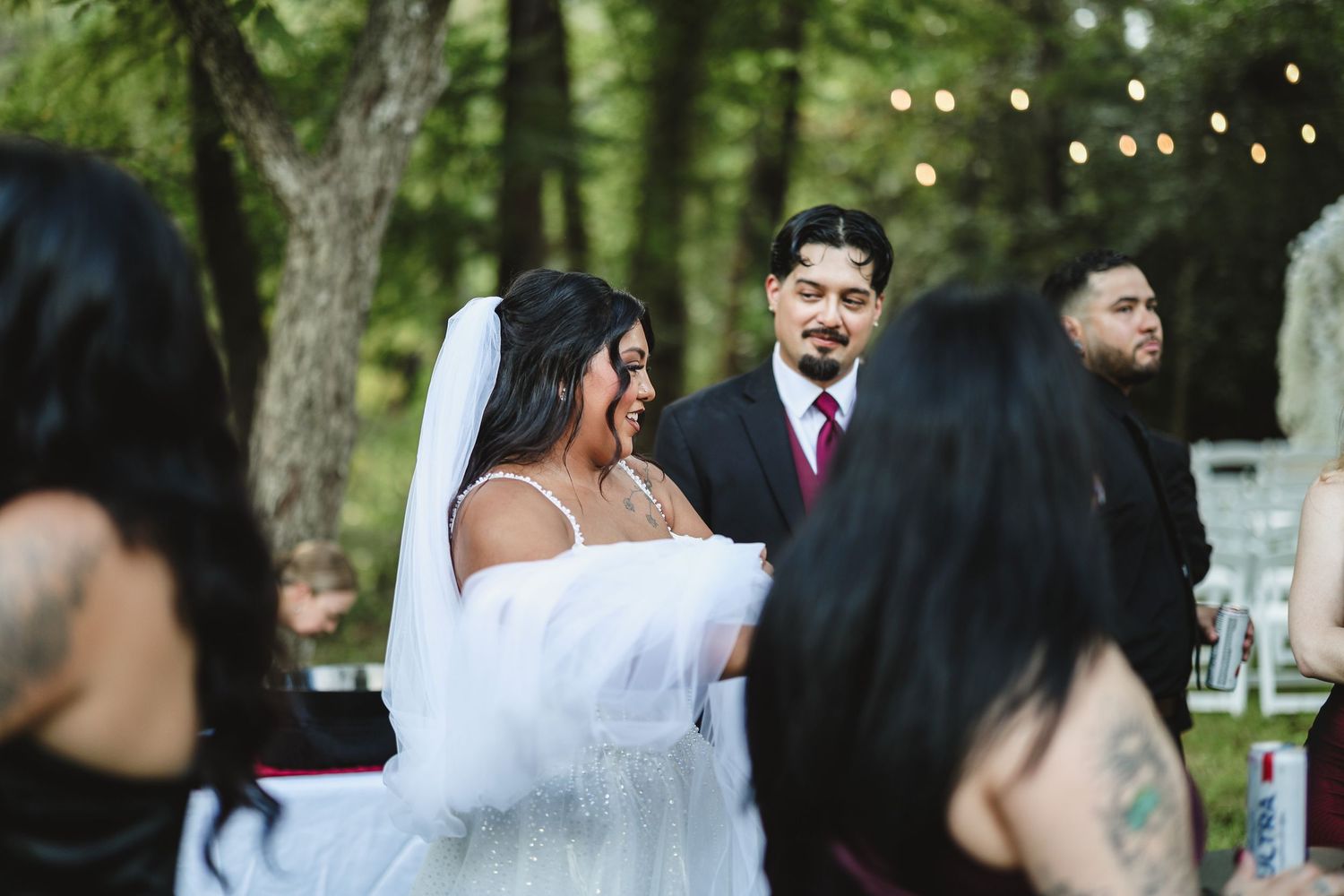 Bride in white gown and veil laughs joyfully with groom in black suit and red tie during outdoor wedding ceremony.