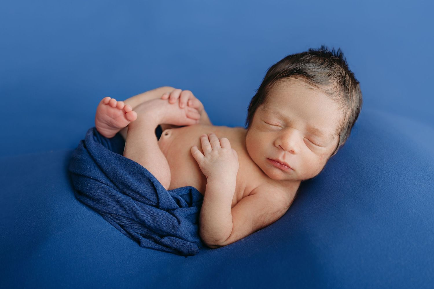 Peaceful sleeping newborn baby curled up on soft blue fabric backdrop in professional studio newborn photography in tampa, florida