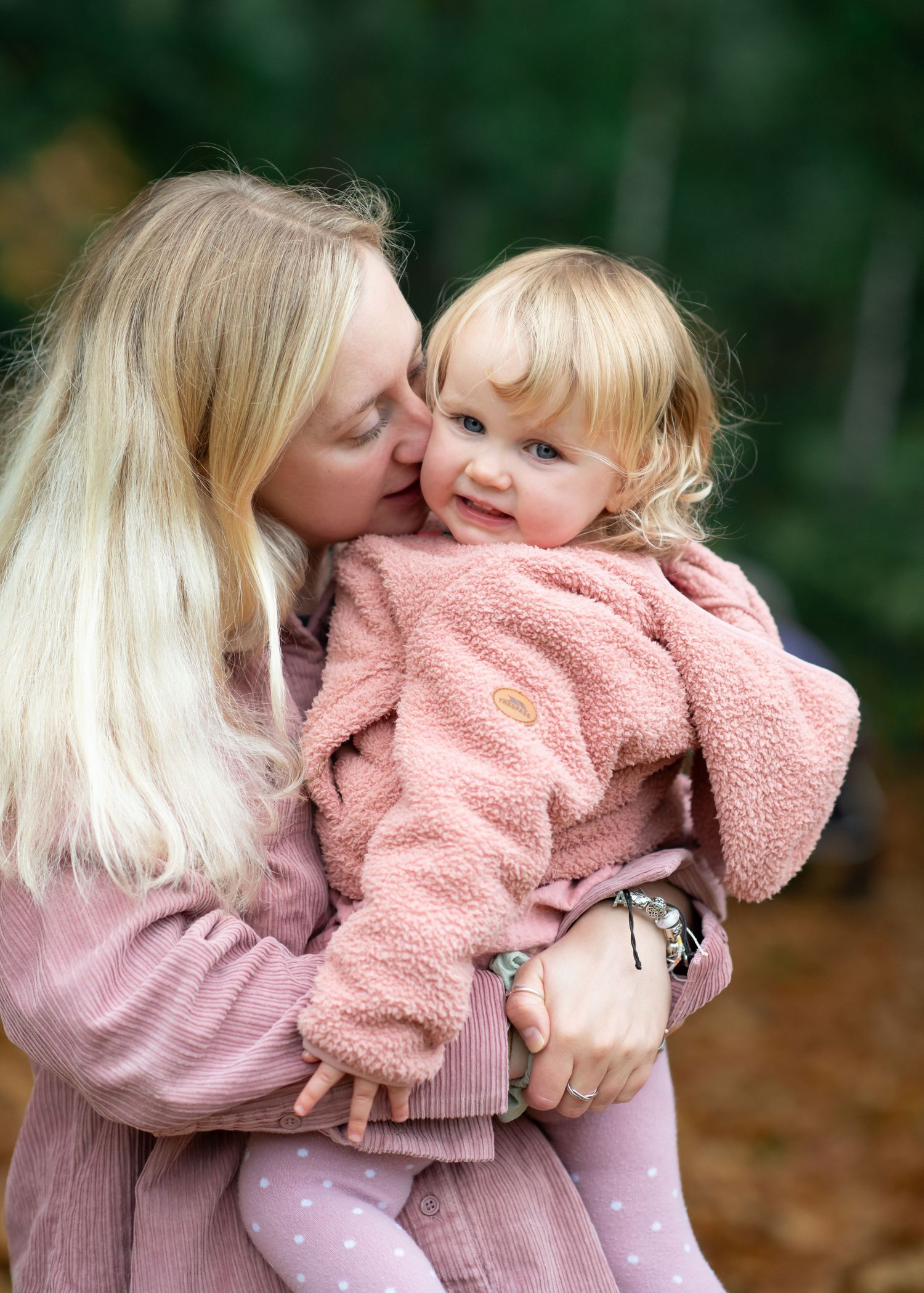A tender moment between mother and daughter wearing matching pink sweaters in an outdoor autumn setting.
