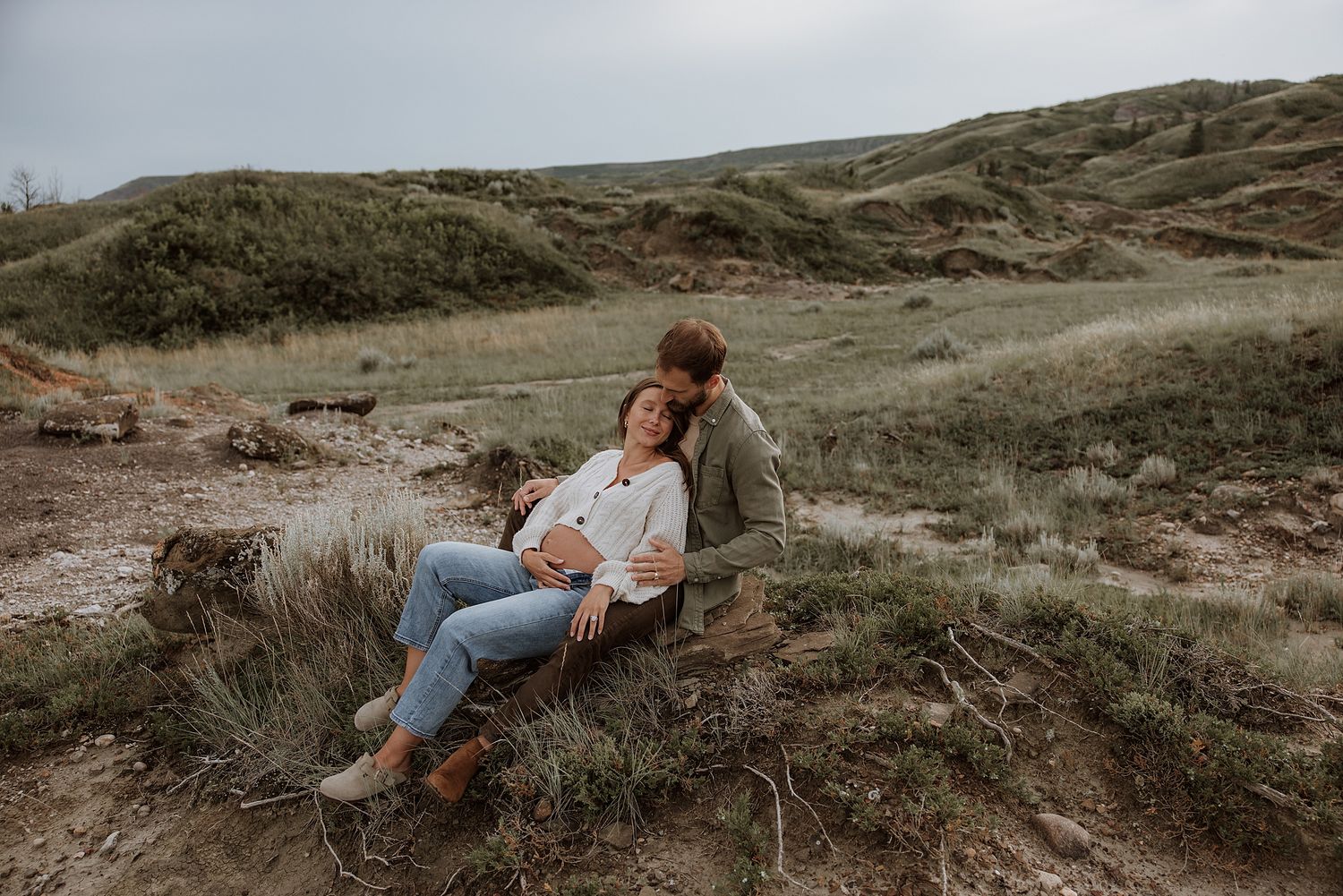 Two people relax together on a grassy hillside overlooking a scenic coastal landscape at sunset.