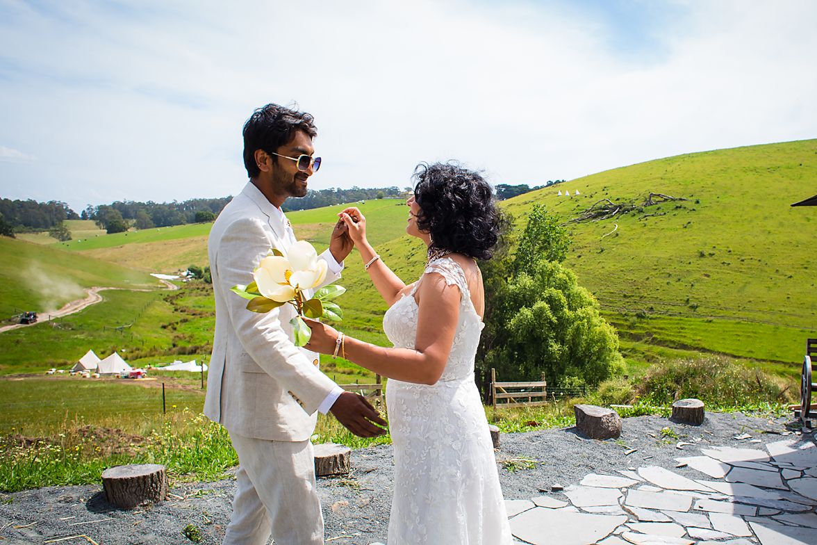 Couple holding hands at outdoor ceremony with scenic green hillside and ocean views in background.