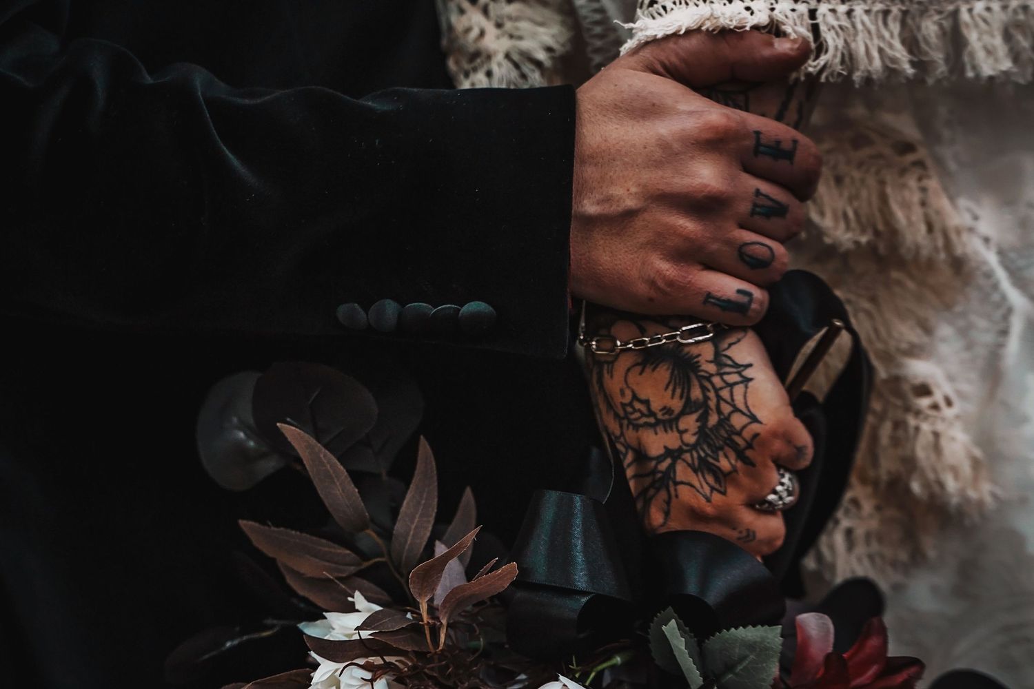 A tattooed hand extends over dark foliage and decorative objects in a moody atmospheric setting.