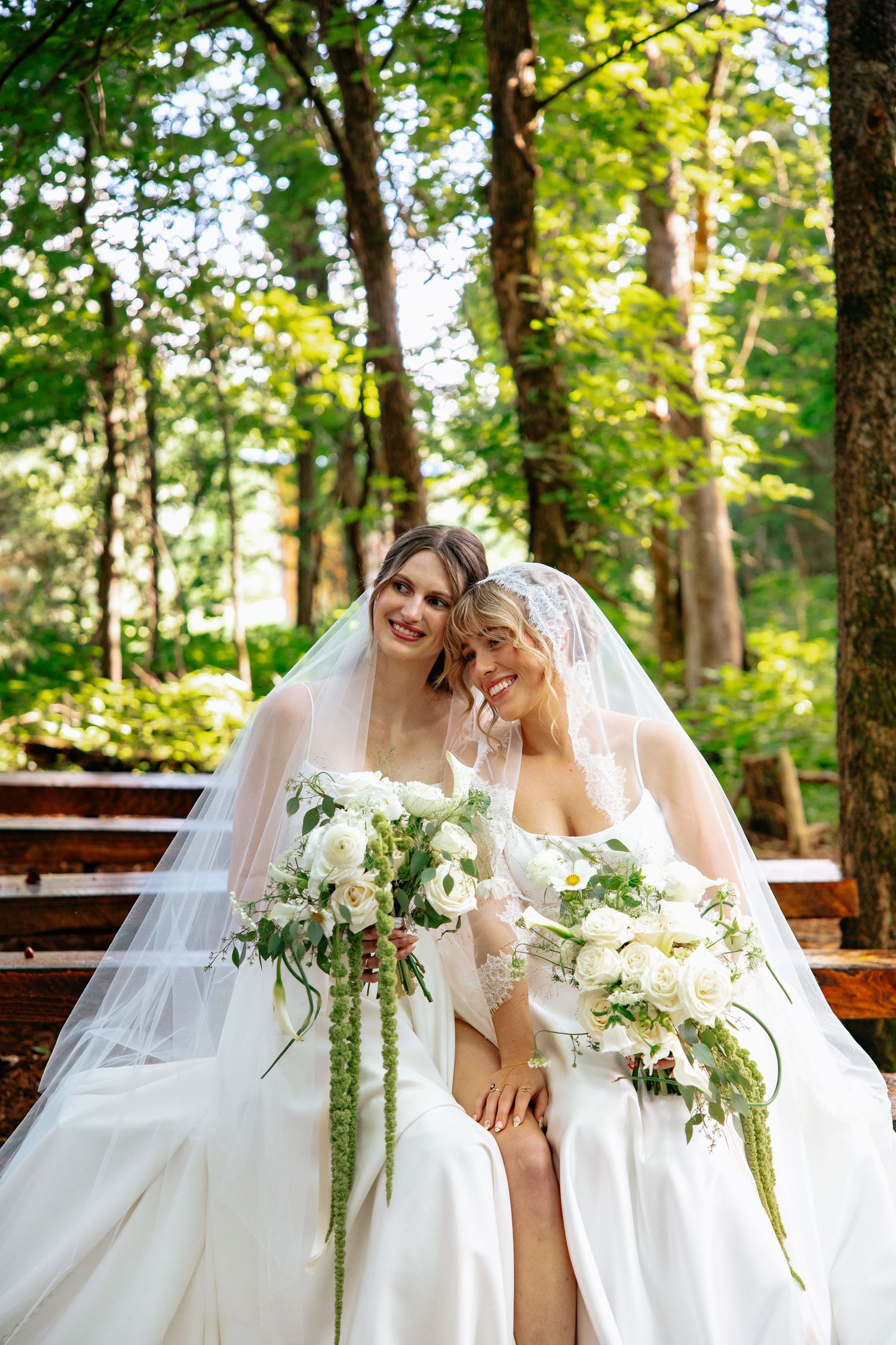 Two brides share a tender moment together in a sunlit forest setting while holding elegant white floral bouquets.