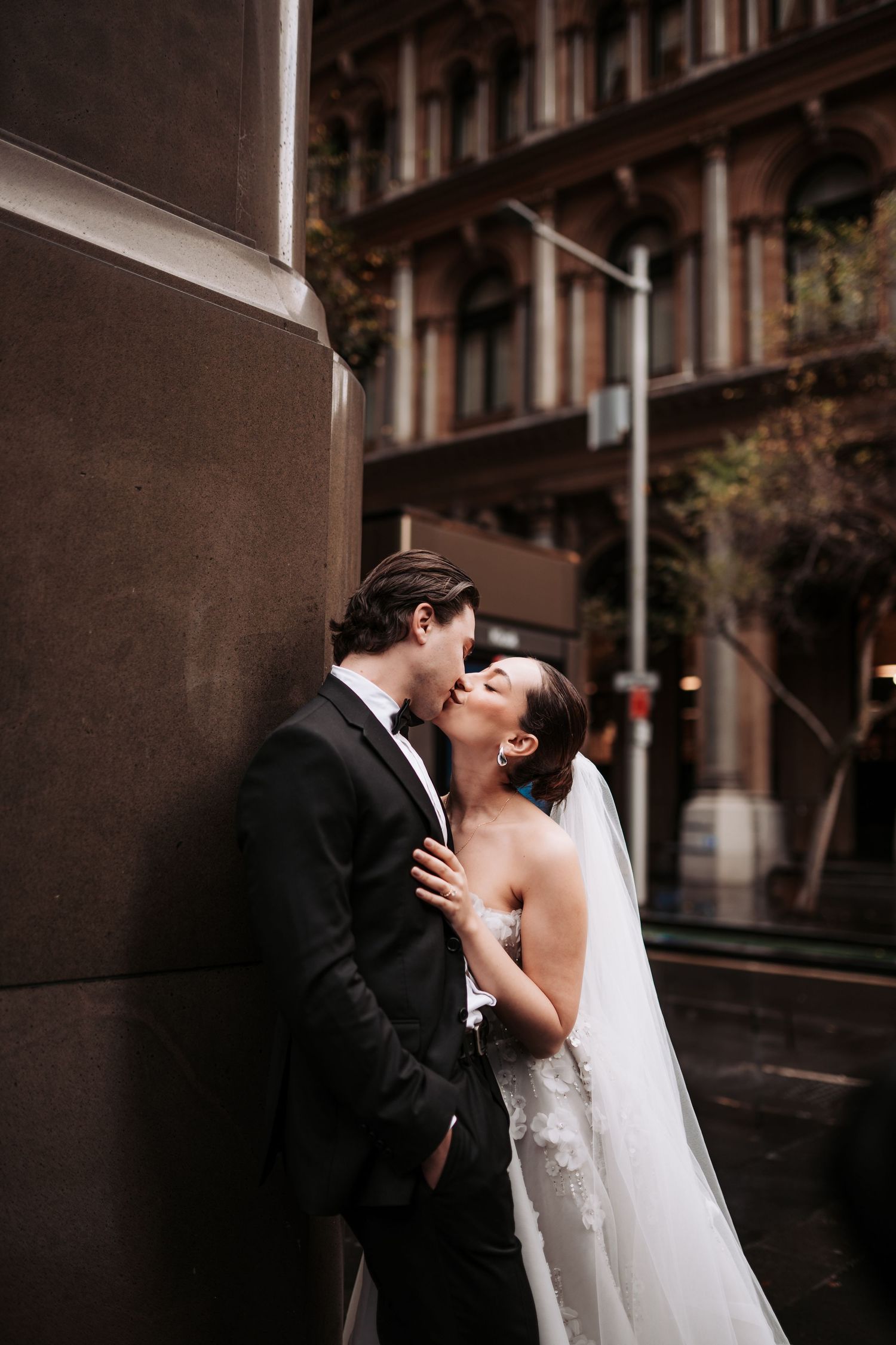 A romantic wedding couple shares a kiss against a brick wall on a city street.